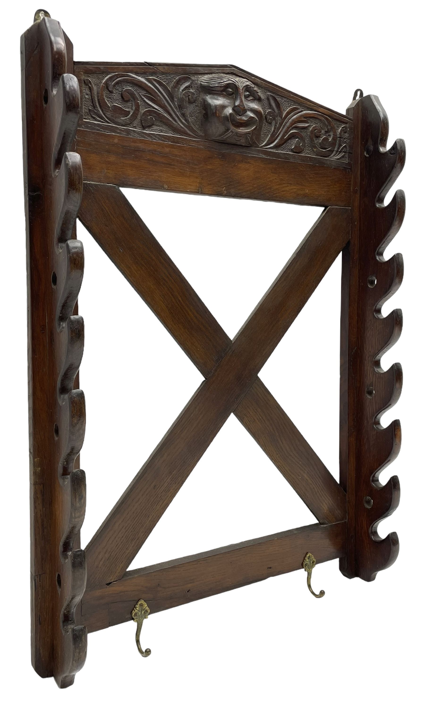 Victorian oak wall hanging whip or stick rack, sloped arch pediment carved with central grotesque mask with extended scrolling foliage, the upright supports cut with rests, x-framed back, two lower hooks