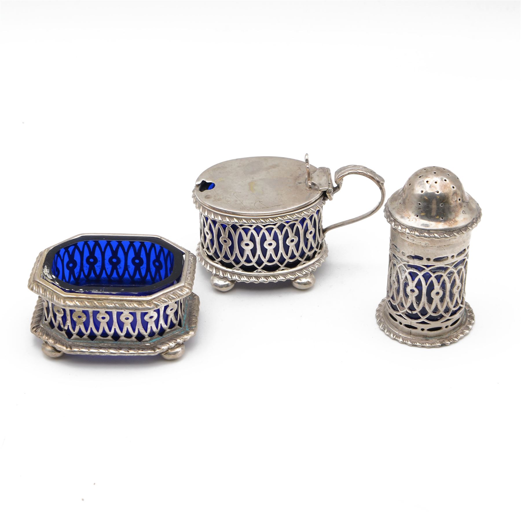Late Victorian silver three piece cruet set, comprising open salt, pepper pot and mustard pot with cover, all with pierced decoration and upon four bun feet, hallmarked John Charles Lowe, London 1899 and 1900, salt with different maker's mark worn and indistinct, all with blue glass liners