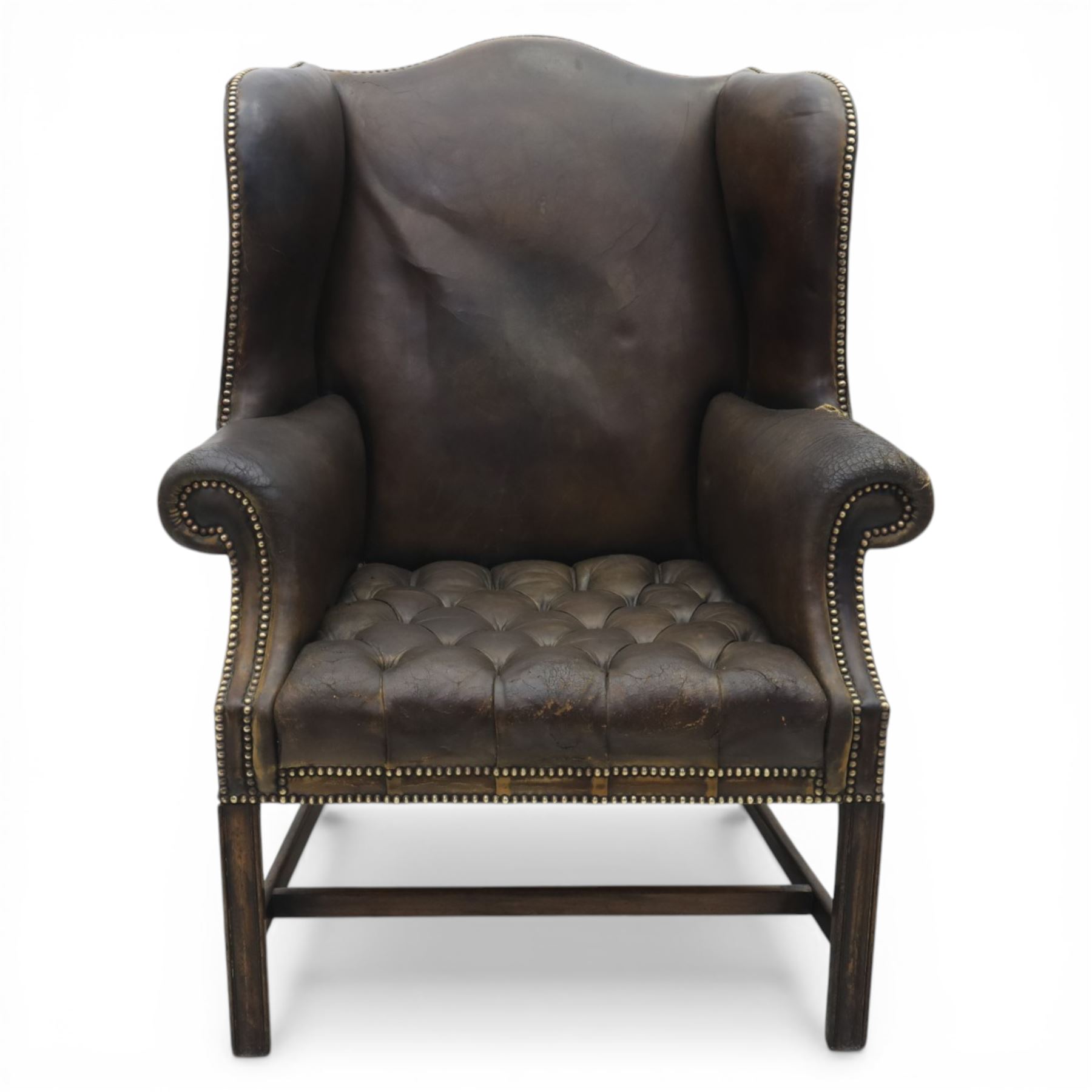 Georgian style leather wing armchair, shaped high back with outscrolled wings above padded arms and deep buttoned seat, close-nailed throughout, raised on square supports united by H-stretcher