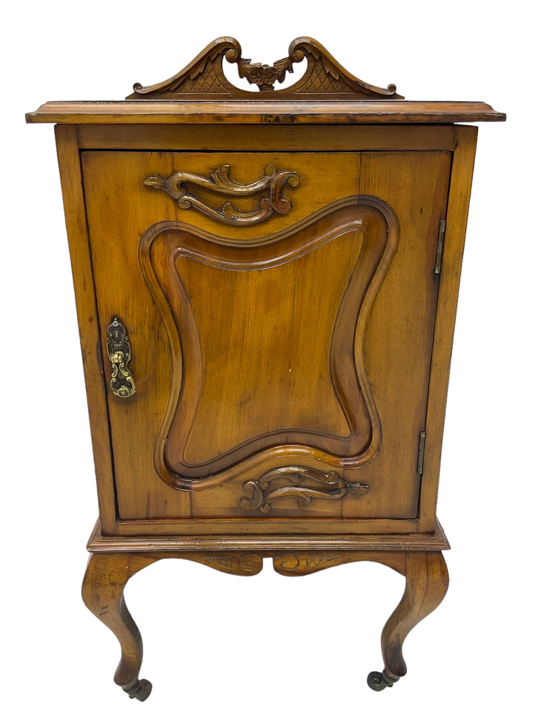 Edwardian Art Nouveau walnut bedside cabinet, raised broken swan neck back with carved flower heads, single fielded panelled door enclosing single shelf, raised on cabriole supports with ceramic castors