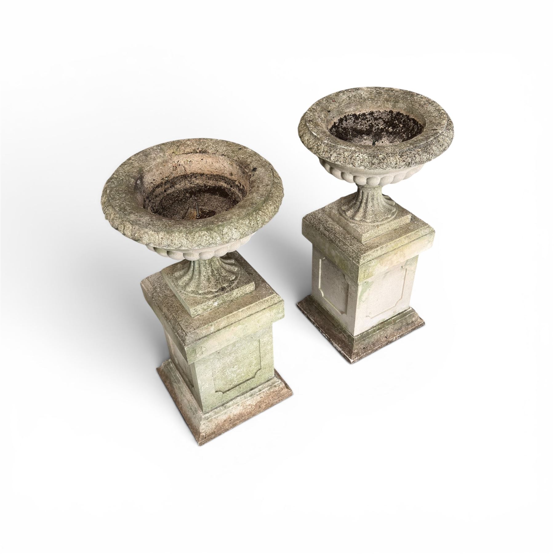 Pair of weathered cast stone Neoclassical design garden Kylix shape urns, foliate moulded rim over a gadrooned underbelly, the scalloped socle over a rectangular pedestal with faux-panelled sides, stamped with stylised ‘H’ monogram