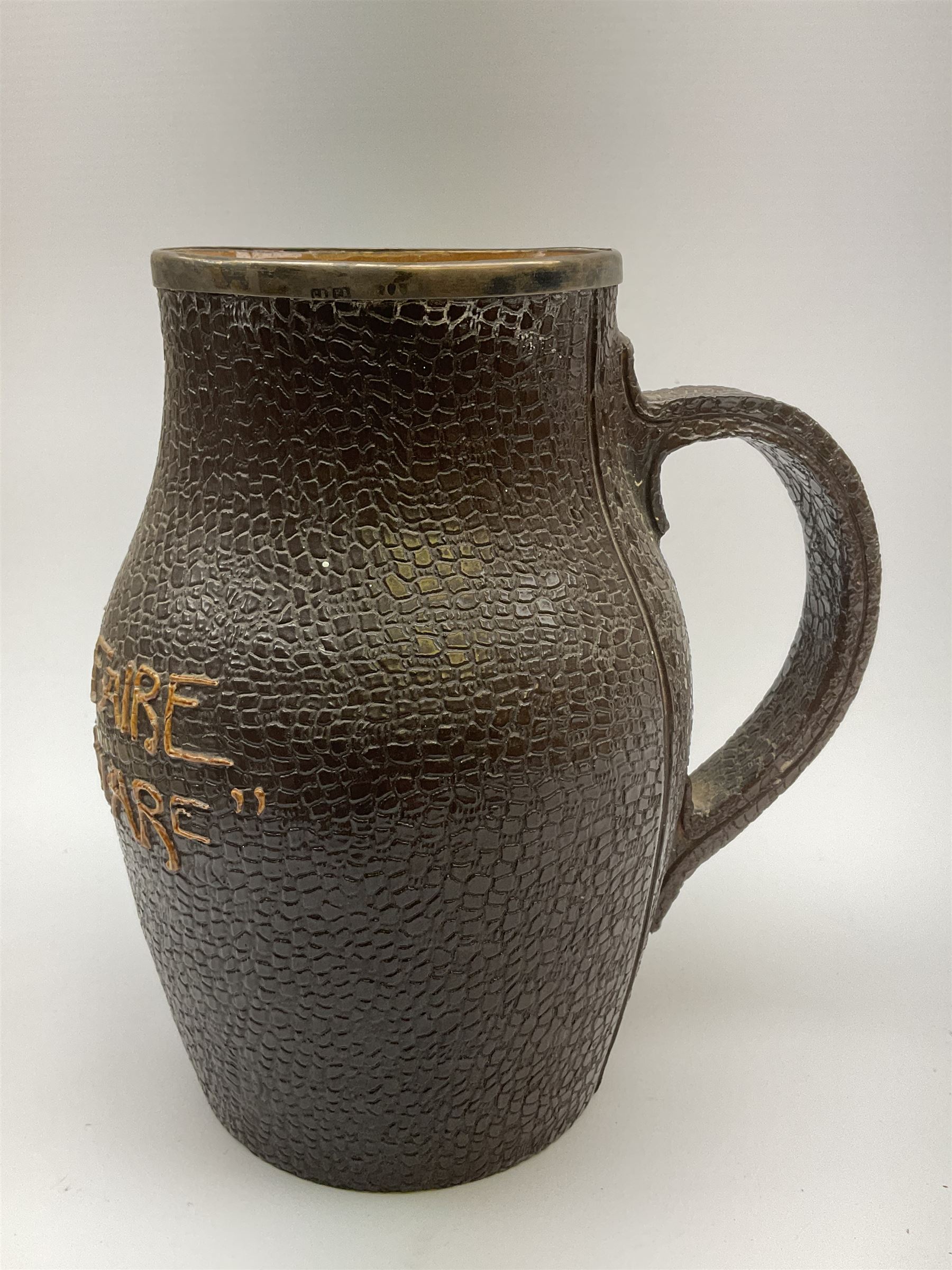 A Doulton Lambeth stoneware jug, with silver mounted collar, hallmarks rubbed, maker's mark probably Cornelius Desormeaux Saunders & James Francis Hollings (Frank) Shepherd, the leather effect body titled 'Drinke Faire Don't Sware', with impressed marks beneath including 'Manufactured for Sidney W Allen 39 White Rock Hastings', H18.5cm. 