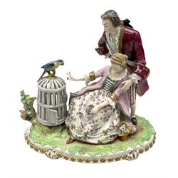 Late 19th/early 20th century Sitzendorf figure group, modelled as a courting couple, the female figure seated offering berries to a parrot stood atop an open bird cage, upon a naturalistically modelled base detailed with encrusted flowers, and further detailed with a gilt foliate band, with blue printed mark beneath, H21.5cm W24cm