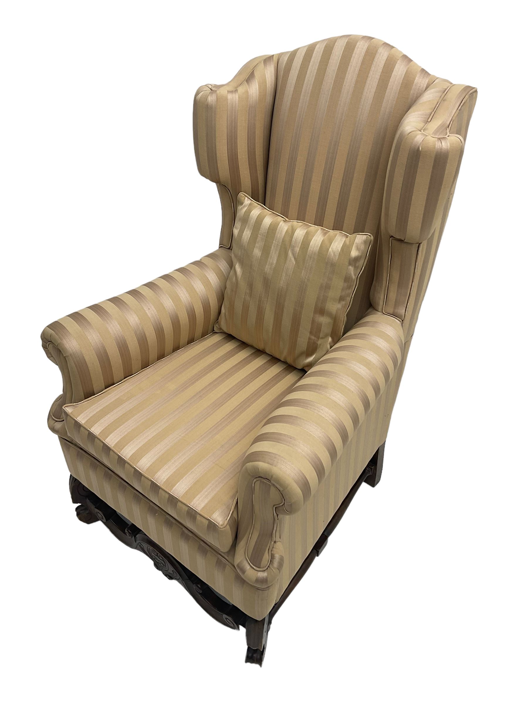 William and Mary style wingback armchair, upholstered in gold striped fabric, on walnut scroll carved supports joined by scroll and foliate carved middle rail, turned H-shaped stretchers, on castors