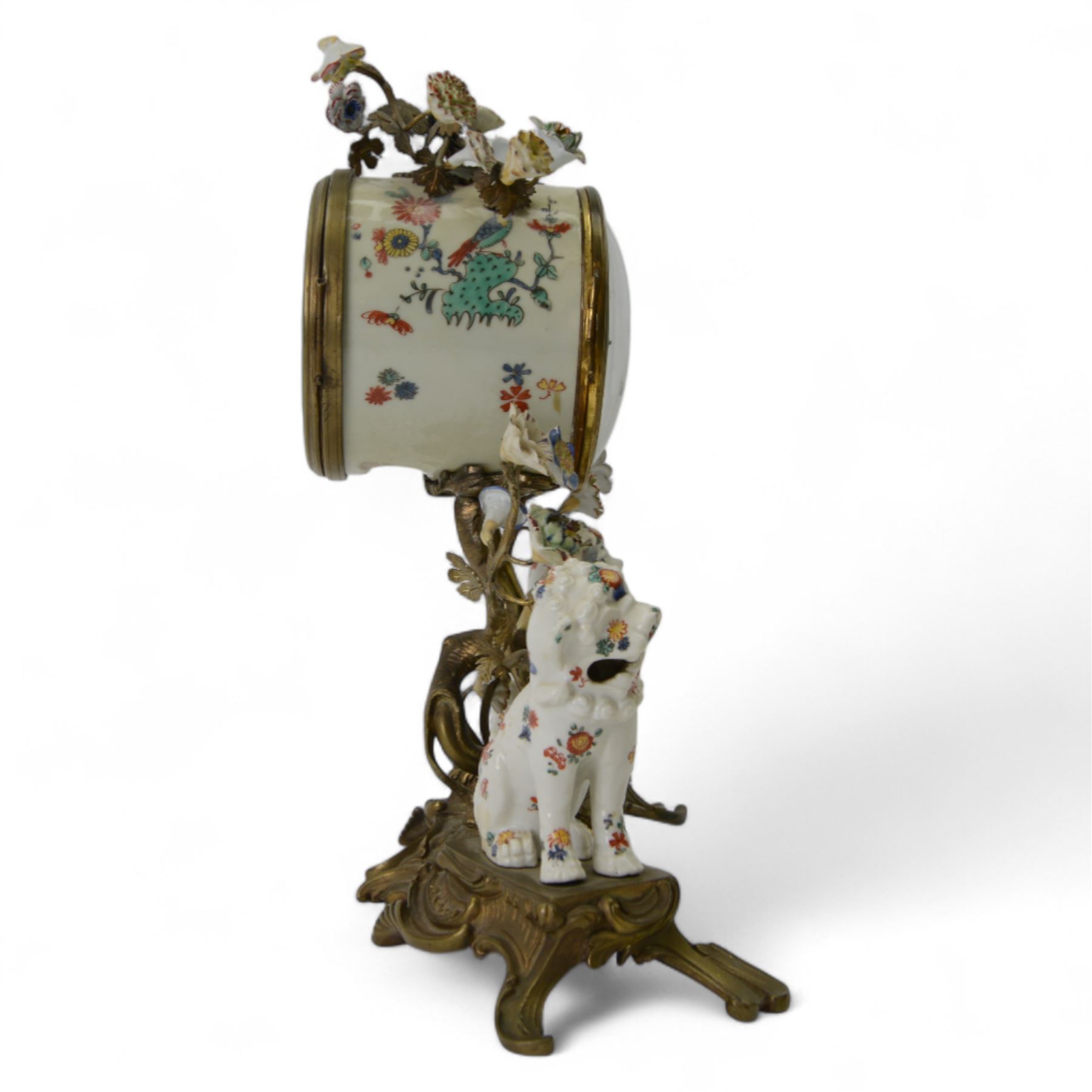 French- early 20th century 8-day Chinoiserie brass and porcelain mantel clock in the 18th century style, brass base with splayed feet profusely decorated with clusters of painted porcelain flowers and two decoratively painted Chinese foo dogs, movement enclosed in a circular porcelain movement case illustrated with birds of paradise and oriental motifs, with a white enamel dial Arabic numerals, minute markers, pierced gilt hands and a floral swag, rack striking Parisian movement striking the hours and half-hours on a bell. With a visible sunburst pendulum and key.