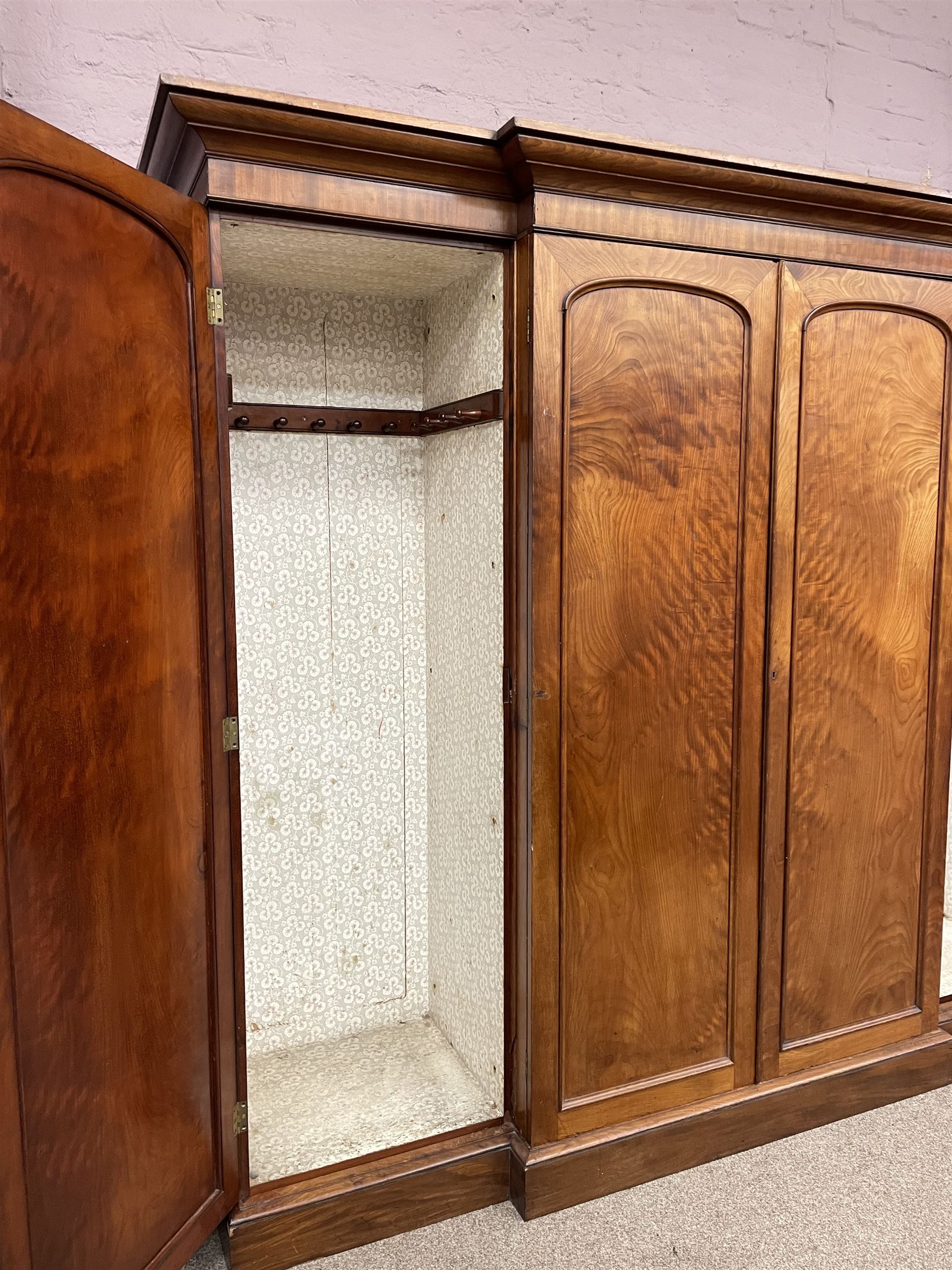 19th century figured mahogany quadruple breakfront press wardrobe, projecting cornice with banded frieze over arch panelled doors, the interior fitted with three sliding trays over two short and three long graduating drawers with recessed brass handles, flanked by two hanging cupboards with hooks