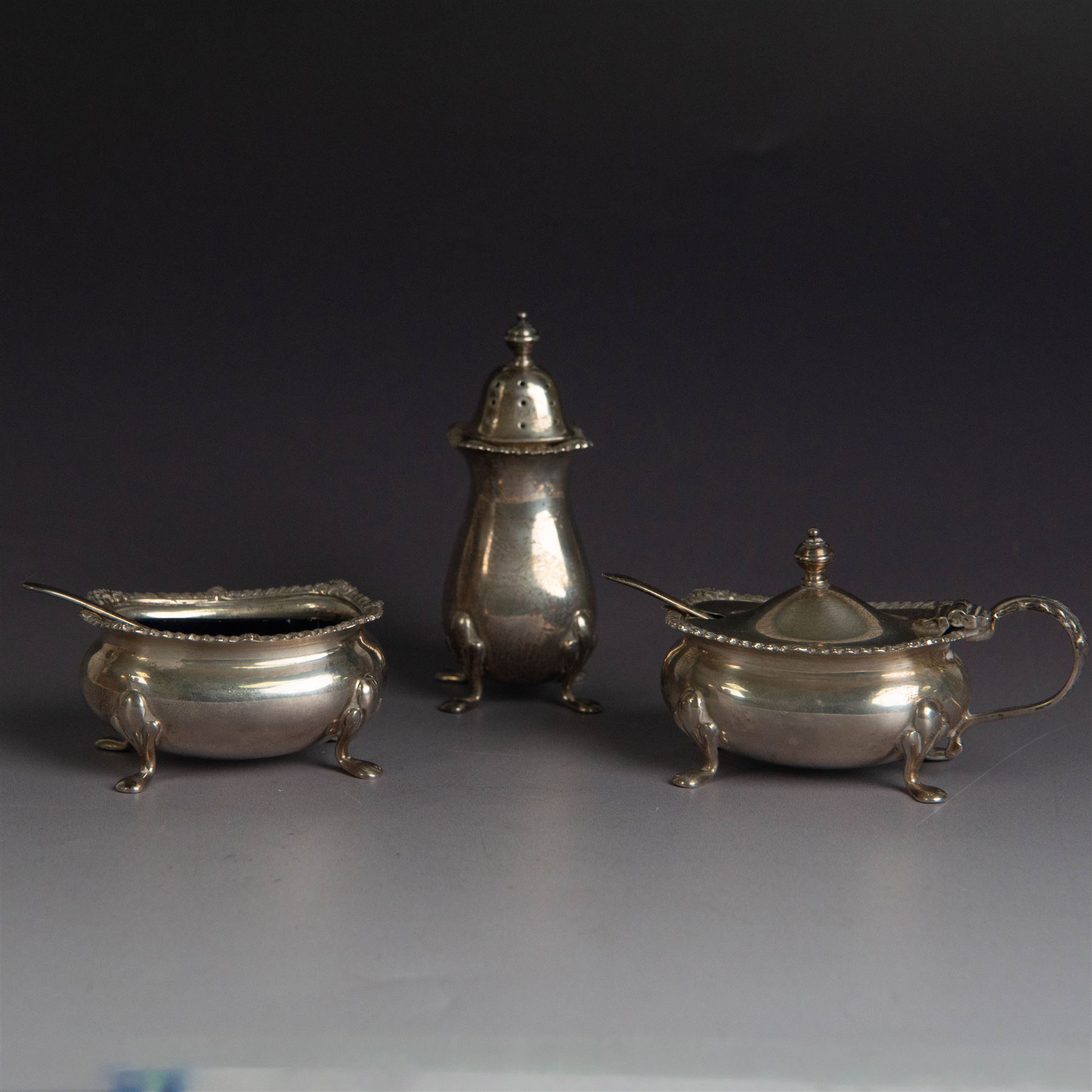 Mid 20th century silver three piece cruet set, comprising pepper shaker, open salt, mustard pot with cover and two condiment spoons, all of oval form with oblique gadrooned rims and upon four paw feet, hallmarked Adie Brothers Ltd, Birmingham 1957, with blue glass liners