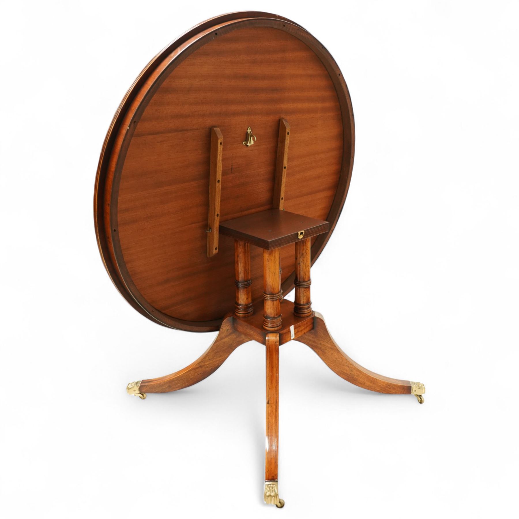 Regency design circular mahogany centre table, the radiating veneered top with contrasting crossbanded edge, on turned column support and four swept legs with brass paw feet and castors