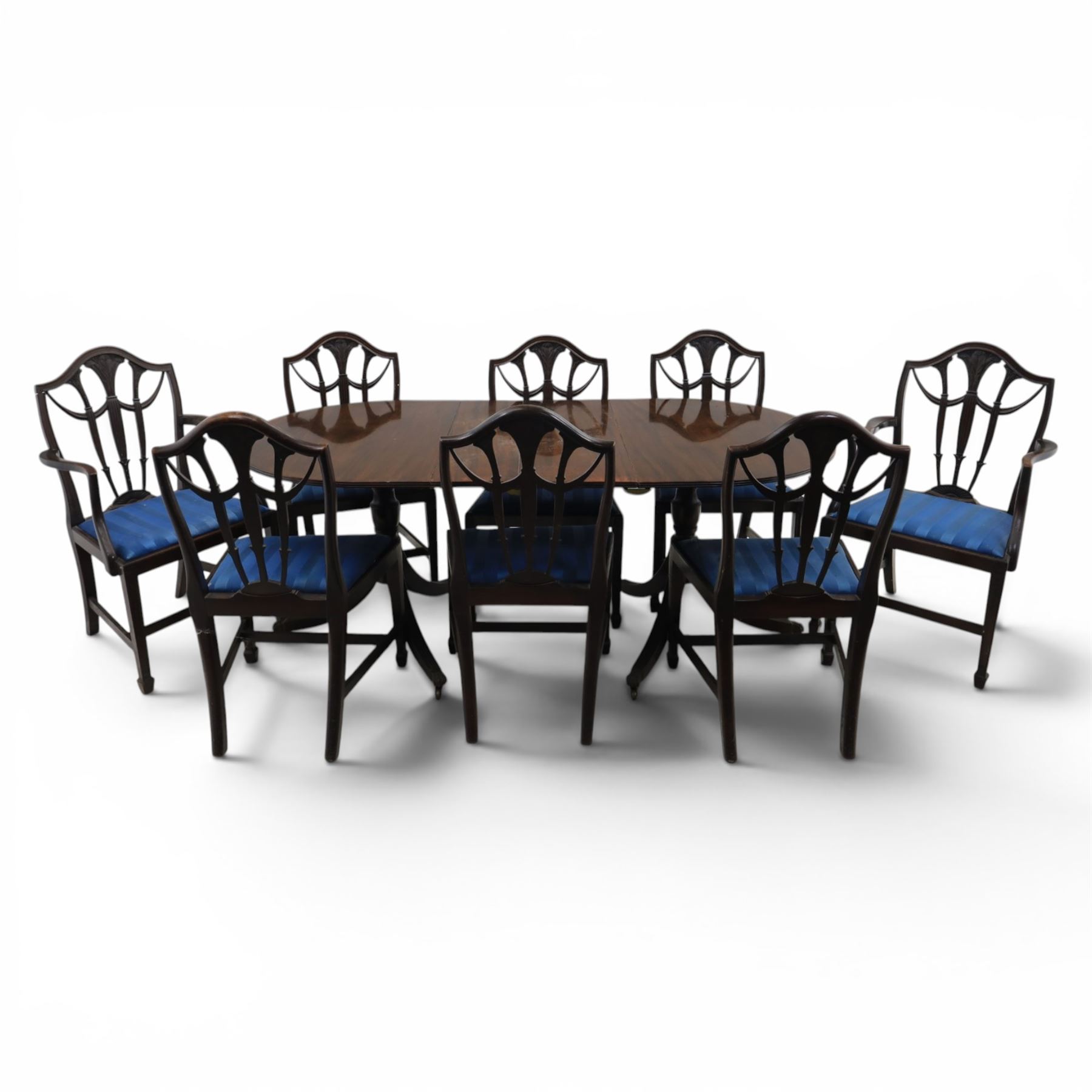 Set of eight George III style mahogany dining chairs, comprising two carvers and six singles, each with shaped top rail above pierced splay carved with feathers, with drop in stuffover seats upholstered in blue striped fabric, raised upon square tapering legs joined by stretchers, together with a mahogany twin pedestal dining table, the oval top with central leaf raised upon turned columns and splayed reeded legs terminating in lion paw feet and brass castors, 