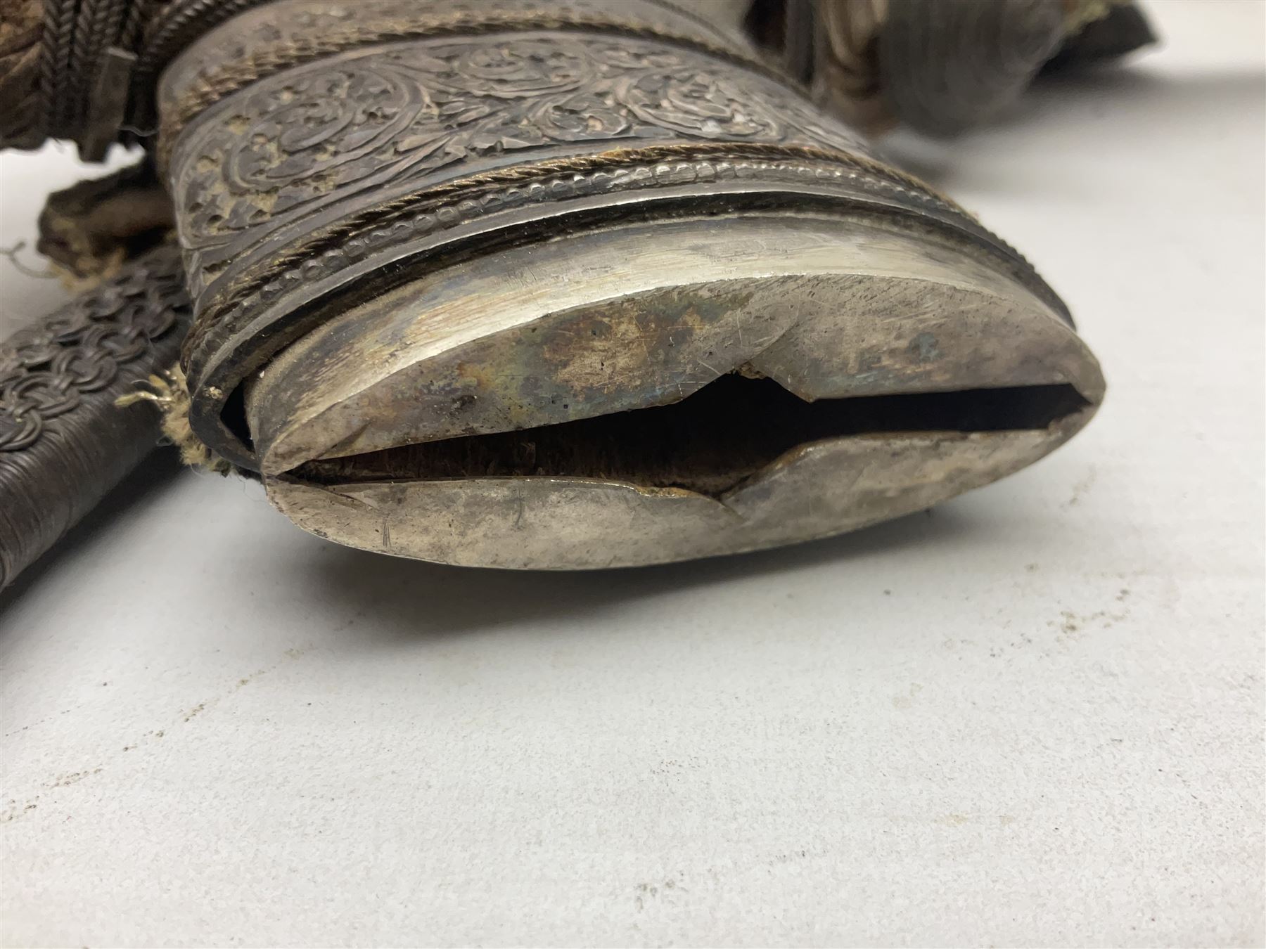 Omani Khanjar dagger, the 19cm curved blade with pronounced medial rib to either side, waisted hilt with dome shaped pommel and silver mounted horn grip decorated with an embossed band; in an intricately decorated silver scabbard with silver rings woven into the belt mount and fitted with original coin mounted silver thread-work belt with additional side knife in sheath verso L34cm overall, belt length 81cm; together with a Nepalese kukri in leather scabbard with one skinning knife (2)