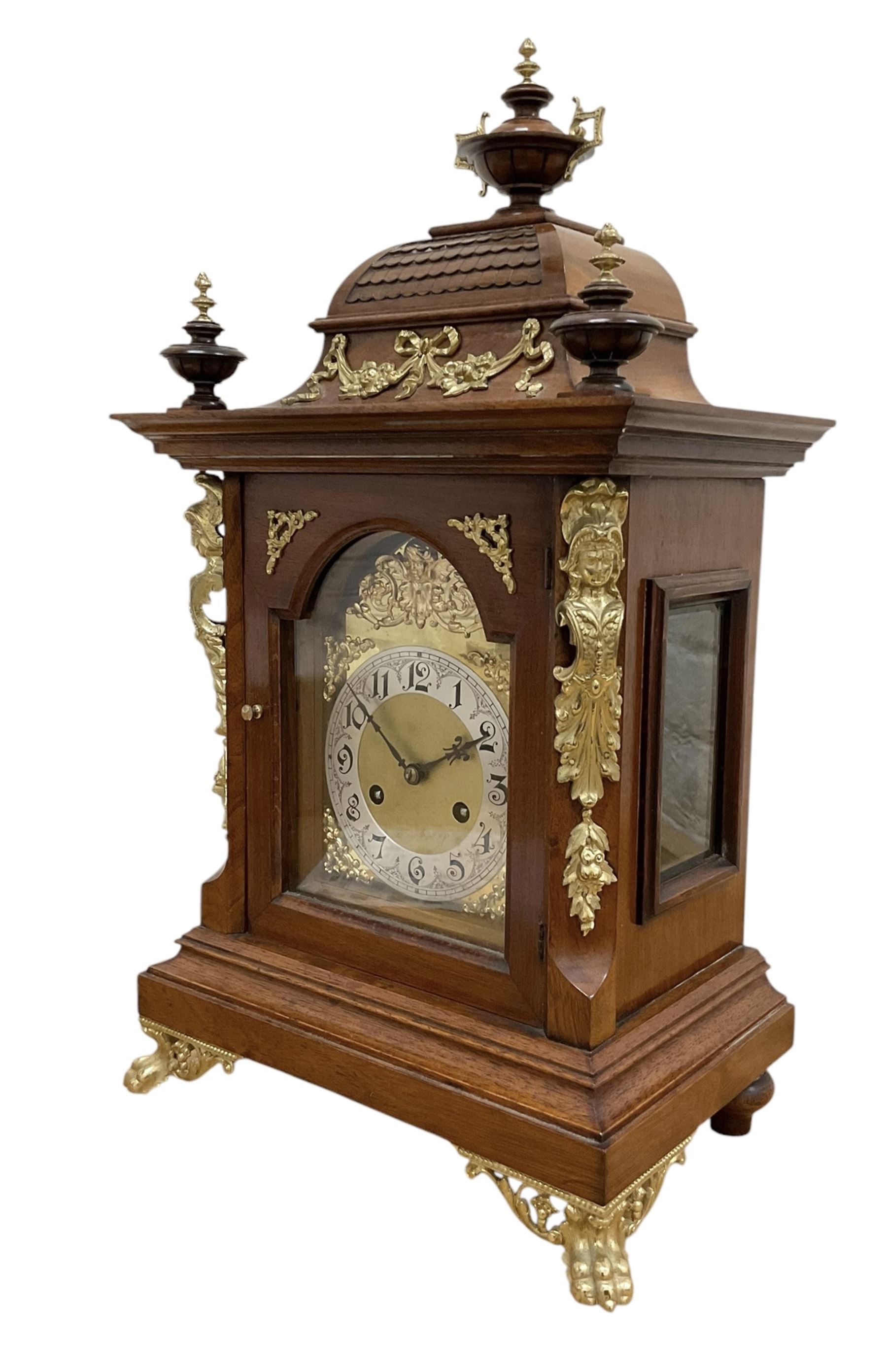 Junghans-German early 20th century walnut cased 8-day mantle clock, with an ogee carved pediment and turned wooden finials, break arch door with brass spandrels beneath flanked by brass caryatids, glazed panels to the sides on a stepped plinth raised on  brass paw feet,  brass dial with a matted centre and silvered chapter with Arabic numerals and Fleur-di Lis steel hands, twin train rack striking movement with a ting tang chime on the quarters. With Key and pendulum. 