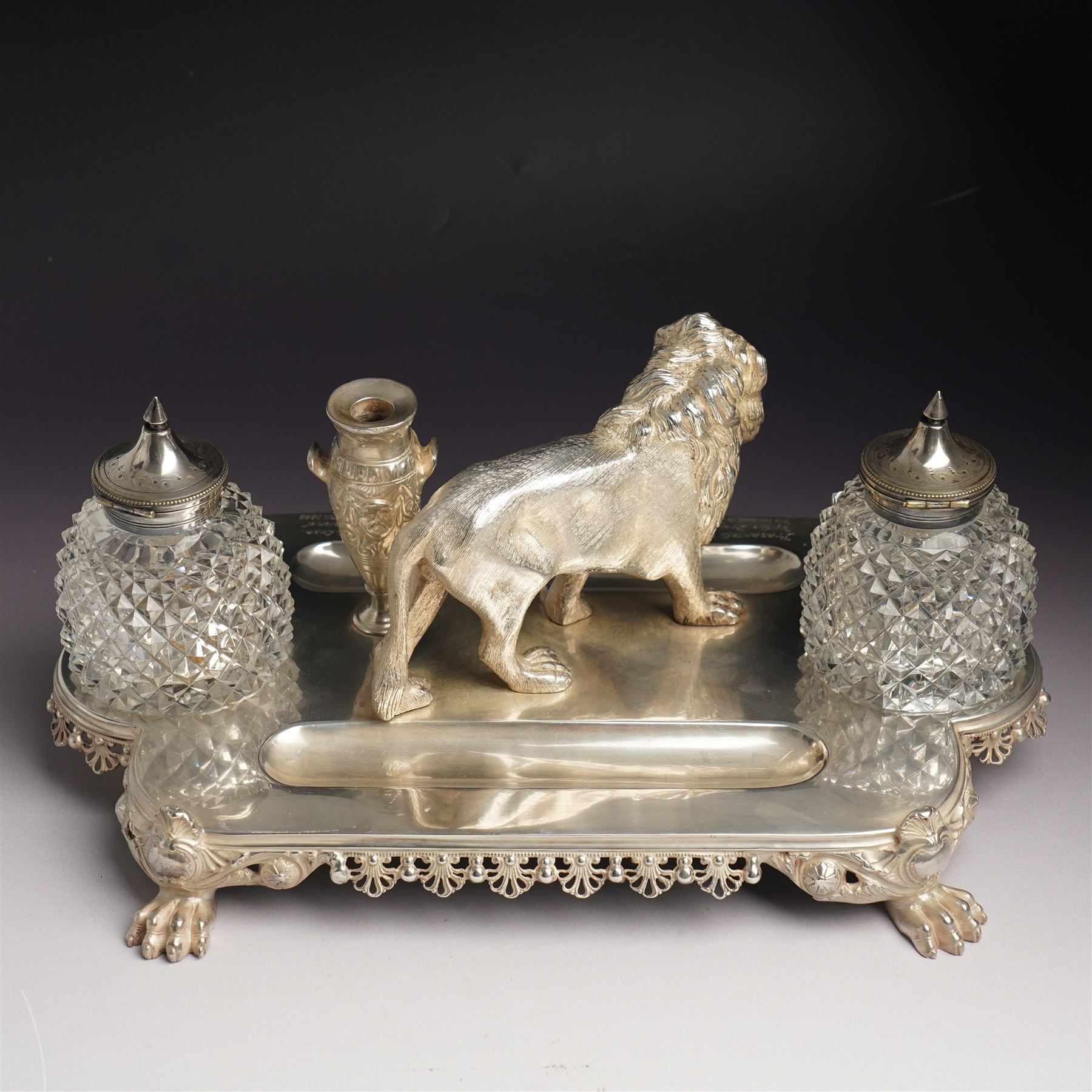 19th century Walker & Hall, silver plated inkstand, oblong and on four paw feet, the border decorated with pierced shell design, each side with a pen-rest, with two cut-glass bottles and central lion and urn finials, engraved to the front with an inscription, H15cm, L30cm