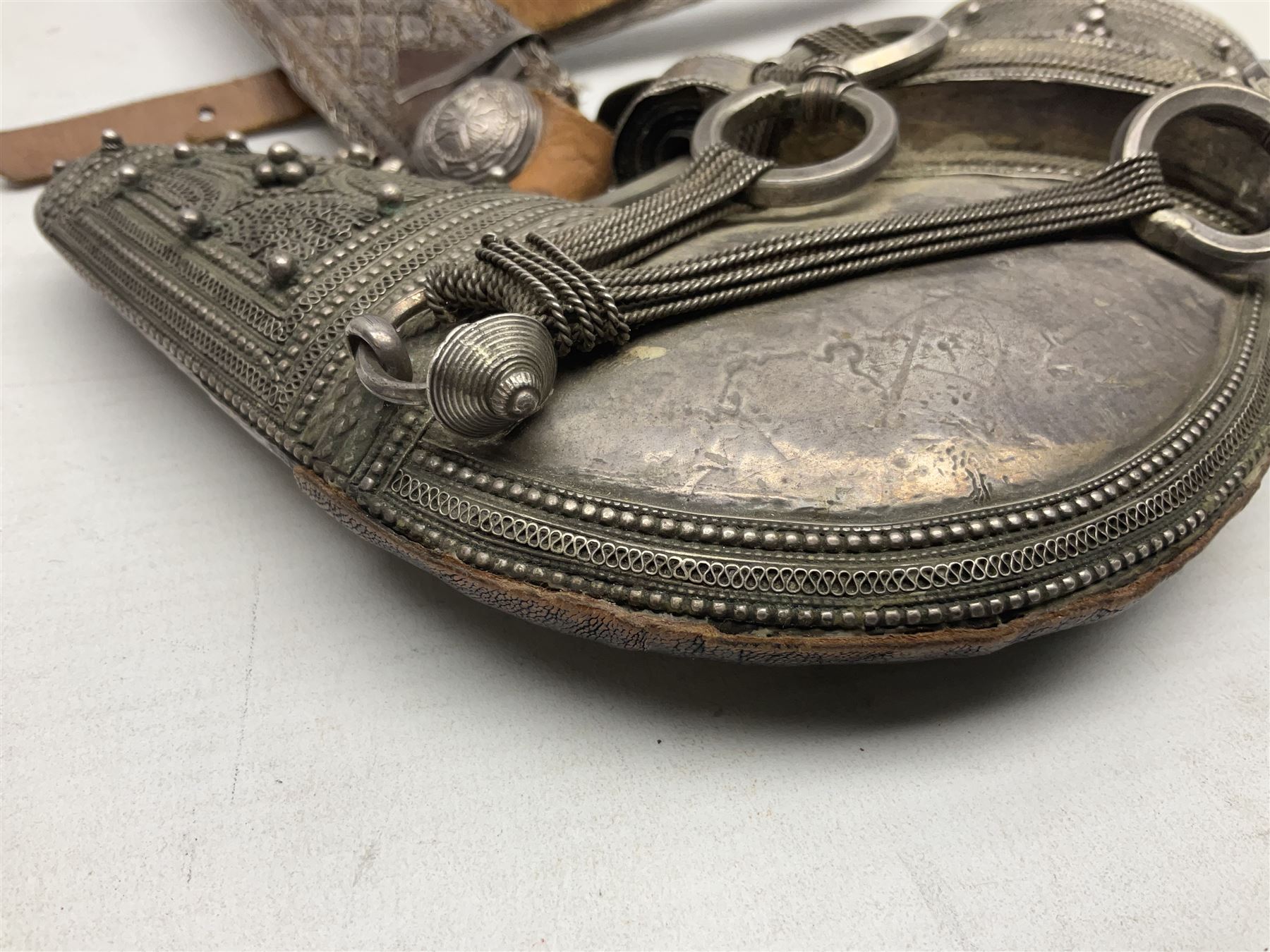 Omani Khanjar dagger, the 19cm curved blade with pronounced medial rib to either side, waisted hilt with dome shaped pommel and silver covered horn grip decorated with filigree bands, floral motifs and beadwork; in an intricately filigree decorated silver and leather scabbard with silver rings woven into the belt mount and fitted with original silver thread-work belt L34cm overall belt length 94cm