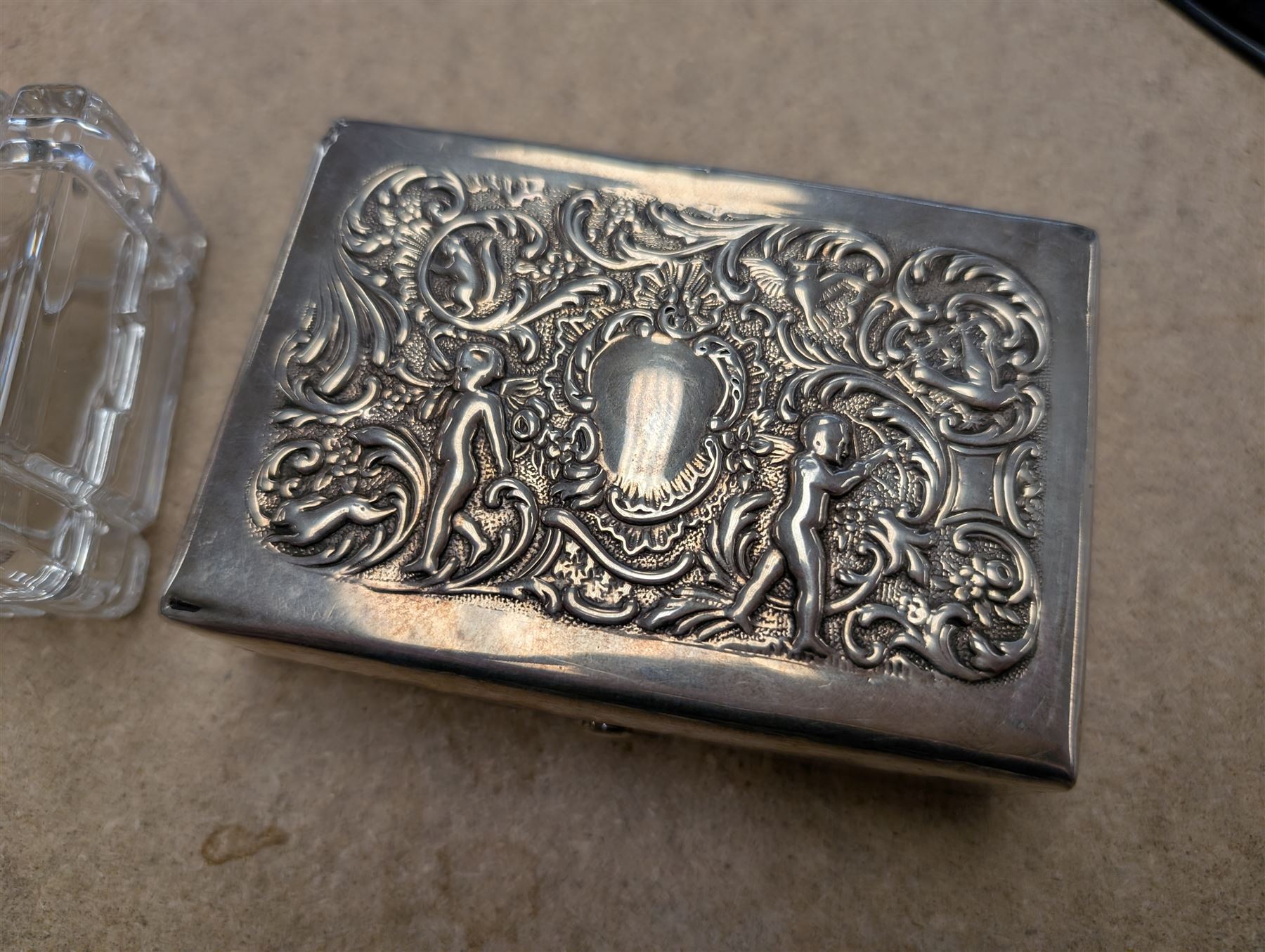 Two silver mounted boxes, including embossed example, decorated with cherubs, birds and scrolls, and a square glass example engraved with initials, both hallmarked 