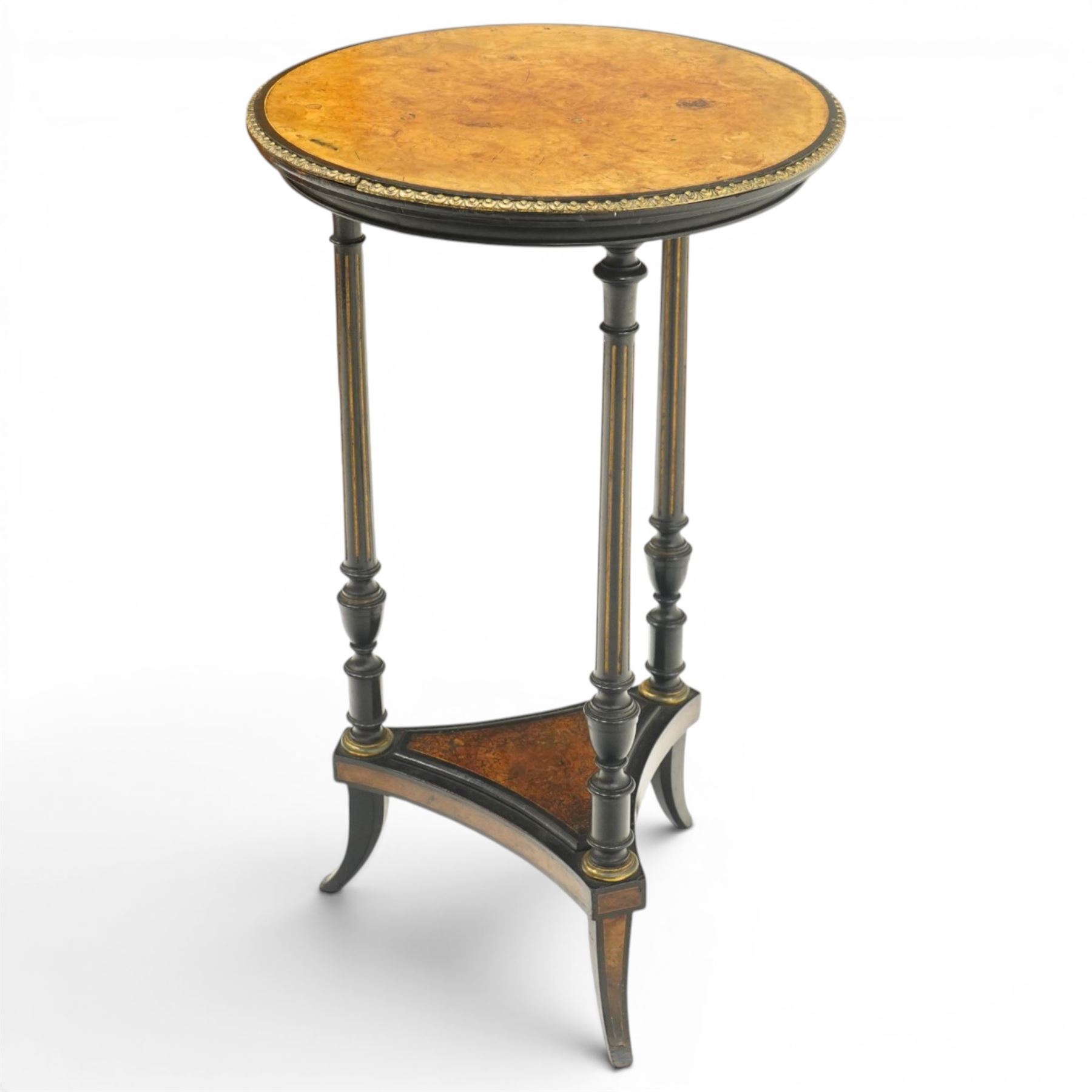 Late Victorian Aesthetic Movement ebonised and figured walnut occasional table, the circular burr top with boxwood strung and ebonised edge, surrounded by gilt metal egg-and-dart moulding, raised on three turned and fluted pedestals with parcel-gilt decoration, united by a triform undertier raised on splayed feet