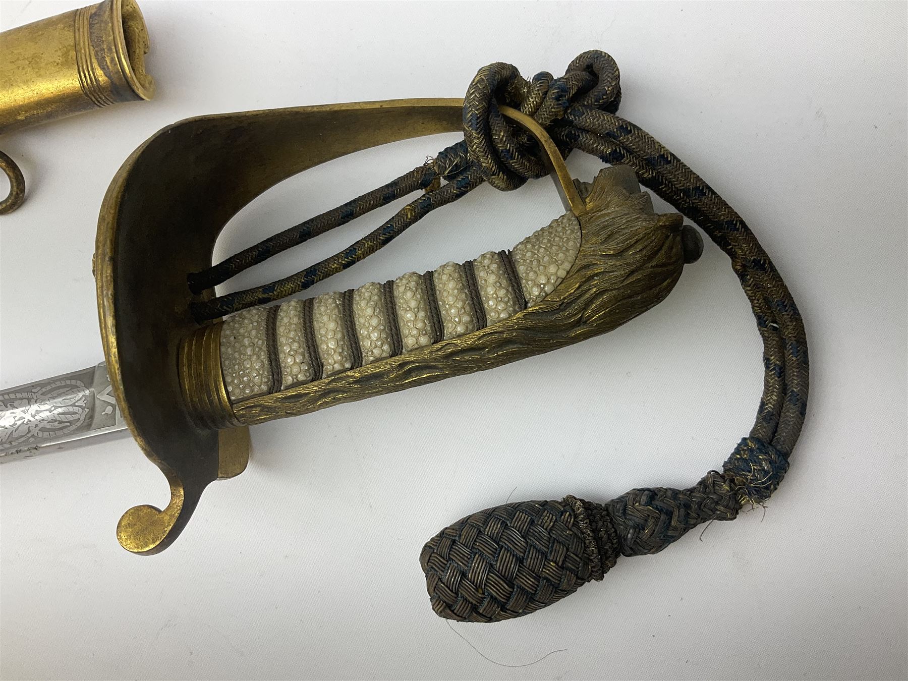 WW1 George V era 1827 Pattern Royal Naval Officer’s sword, the 80cm fullered steel blade decorated with crown, fouled anchor etc and marked Gieves Portsmouth London Edinburgh Devonport to the ricasso, gilded hilt with fouled anchor and crown, wire-bound ray skin grip, lion’s head pommel with mane backstrap, folding/locking langet and full bullion knot; in  leather scabbard having twin brass lockets with hand engraved panels and twin suspension rings L94cm overall. Provenance: By direct family descent from Brian Heathcote HMS Conqueror later Superintendant in the Liverpool Special Constabulary.