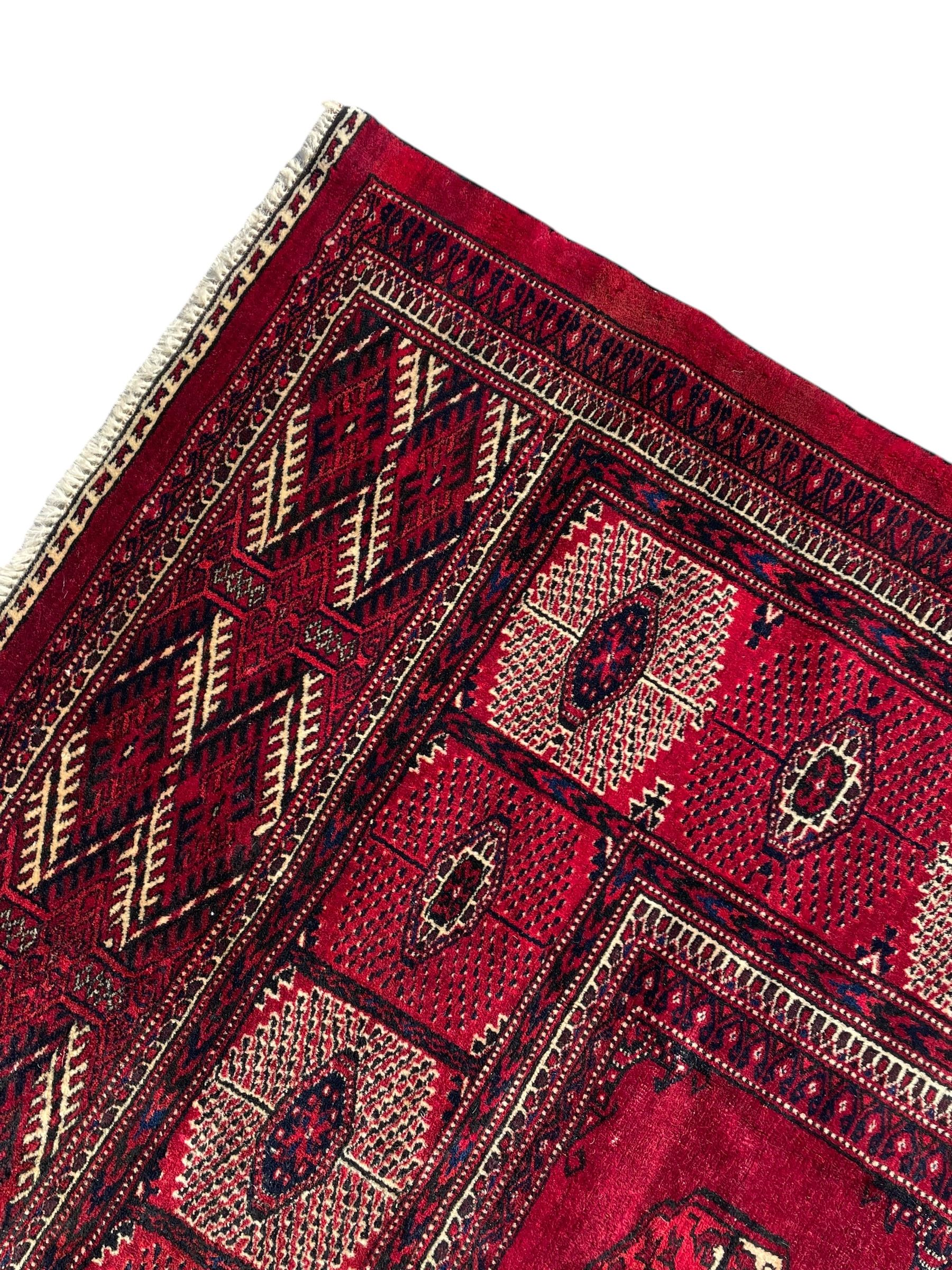 Afghan Bokhara red ground carpet, the field decorated with four rows of Gul motifs interspersed with Hac motifs, the border with lattice pattern panels within small geometric guard stripes, geometric patterned end panels