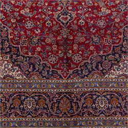 Central Persian Kashan hand-knotted carpet, madder red field with central cusped medallion in navy and ivory enclosing a floral rosette and radiating scrolling arabesques and palmettes, across a densely ornamented ground of finely drawn vinery enriched with a profusion of flowering sprays and stylized blossoms, spandrels in deep indigo and principal border of navy ground decorated with a continuous palmette and vine-scroll flanked by multiple guard stripes, with cotton fringe ends