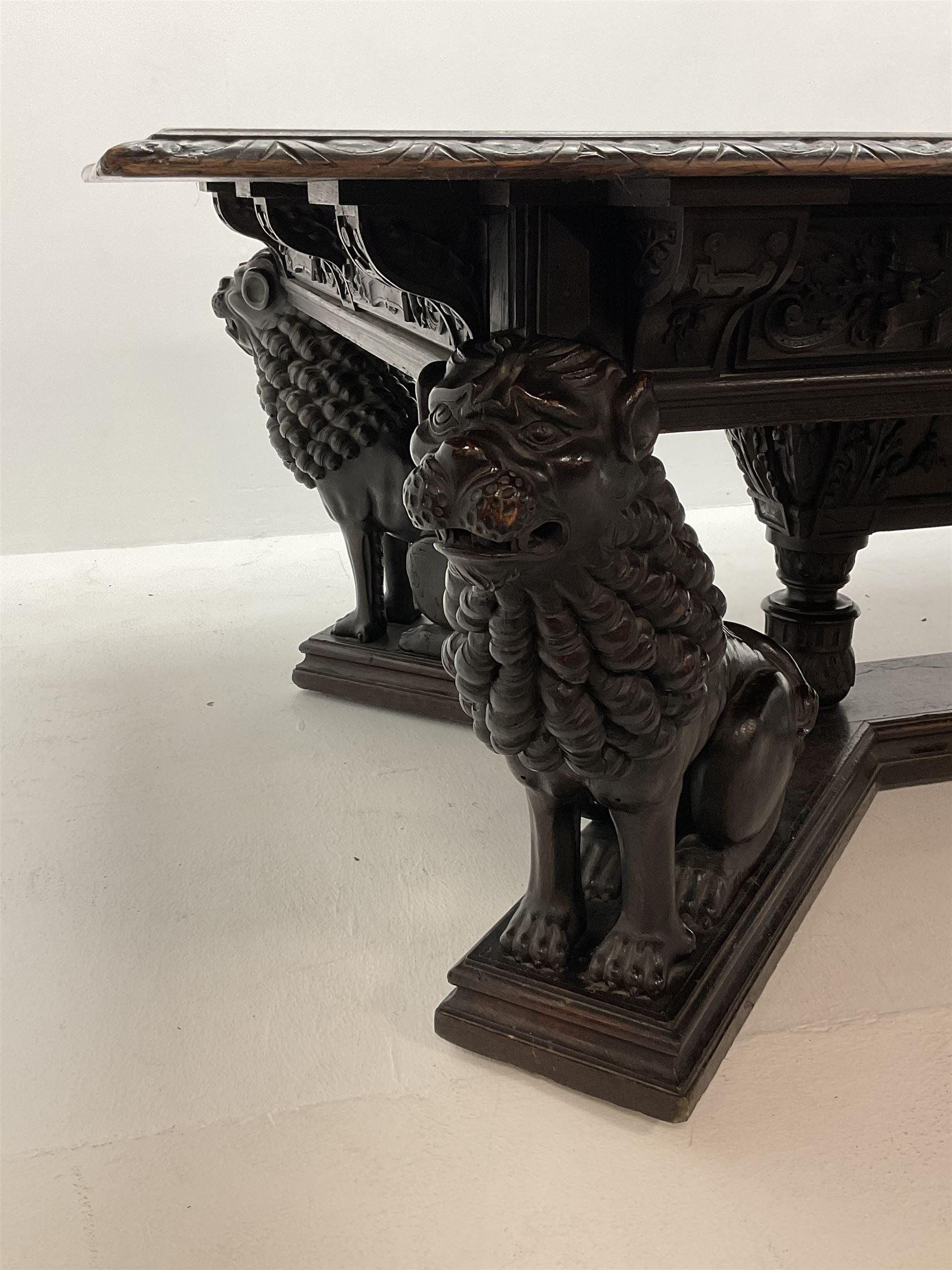 Large 19th century carved oak library table, the rectangular top with lunette carved edge over cartouche panelled and carved  scroll frieze, the central tapering rectangular support relief carved with masks and garlands, on four carved seated lion supports at each corner, on flat angular stretcher with recessed castors stamped ‘Bartholomew & Fletcher, London’