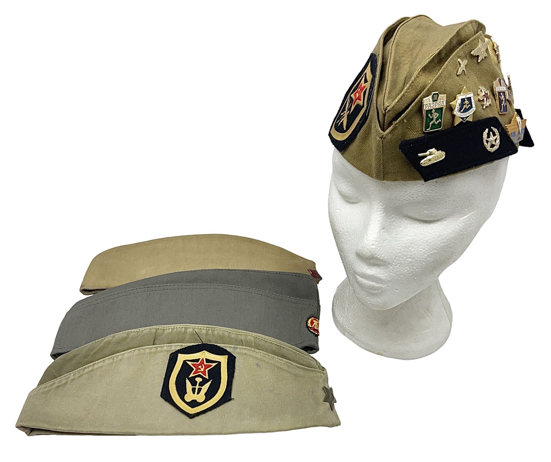 Four side caps - WW2 Russian, post-WW2 Russian and two East German; all with badges, one with multiple metal badges (4)