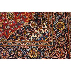 Central Persian Kashan hand-knotted rug, madder red field, with central lobed medallion with pendants, issuing an all-over design of scrolling vine, palmettes, and floral sprays, with conforming spandrels, enclosed by a broad indigo palmette and vine border framed by guard stripes in a palette of navy, blue and ivory tones, with cotton fringe ends 