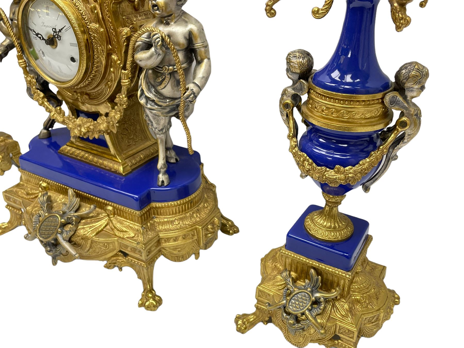 A 20th century continental gilt metal Lyre mantle clock with a pair of matching six light candelabra, spring driven movement housed in a gilt drum case on a brass and cobalt blue base with paw feet, two bronzed metal figures of mythological cherubs with animal legs and cloven hooves supporting a festooned garland swag, eight-day twin barrel striking movement with a floating lever balance escapement, striking the hours and half-hours on two bells, white enamel dial with roman numerals, minute track and pierced steel hands, dial inscribed “Imperial”. 
