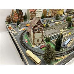Table top 'N' gauge continental scenic layout as a split level town with various loops of track, station with numerous platforms, engine shed, assorted buildings, roads with motor vehicles and figures, tunnels, trees, river with bridge, tram depot etc 122 x 52cm