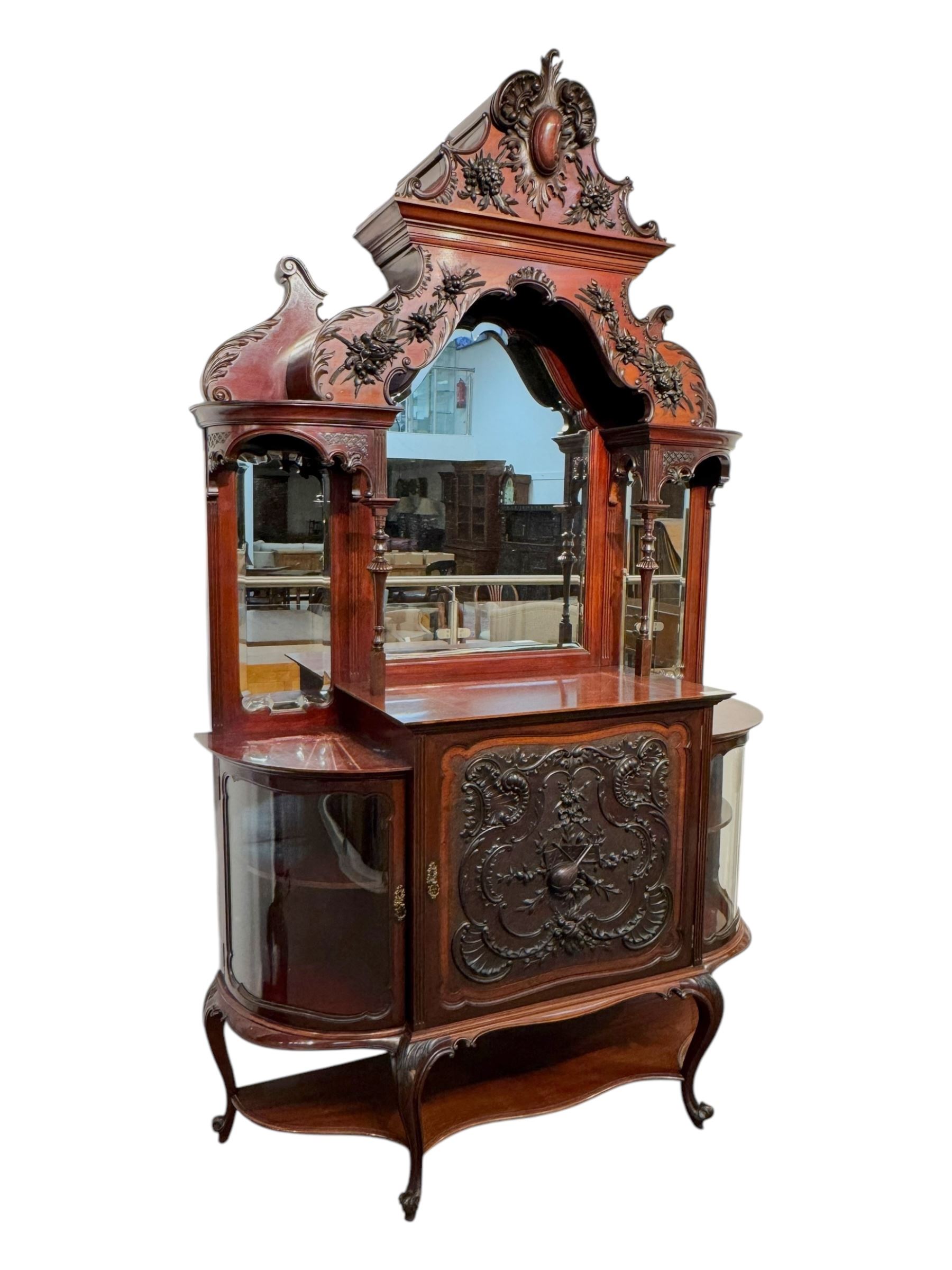 Late 19th century mahogany mirror back cabinet, shaped C-scroll pediment decorated with carved acanthus leaf cartouche and shells, shaped canopy top with applied foliate carvings over triple bevelled mirror back, central cupboard enclosed by panelled door carved with musical instruments, scrolls and shells, flanked by two glazed cabinets, on acanthus leaf carved cabriole supports united by undertier 
