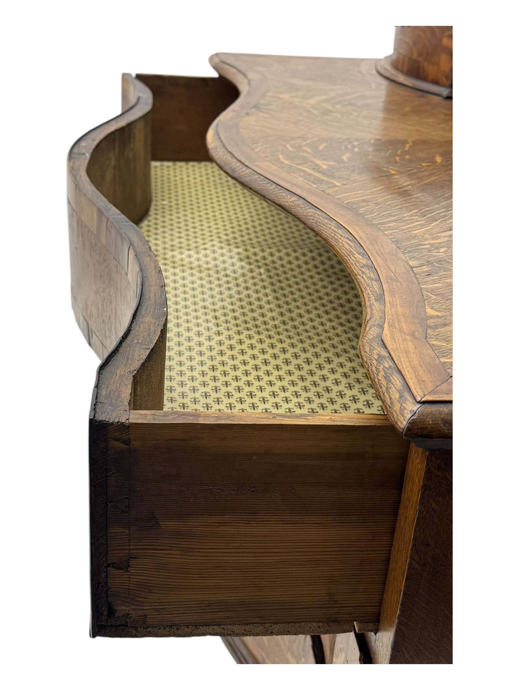 Late 19th century continental oak commode, serpentine fronted with parquetry and crossbanded decoration, the raised superstructure back fitted with a central bank of five recessed drawers flanked by concave drawers, above three full-width drawers with brass ring handles, raised on cabriole supports