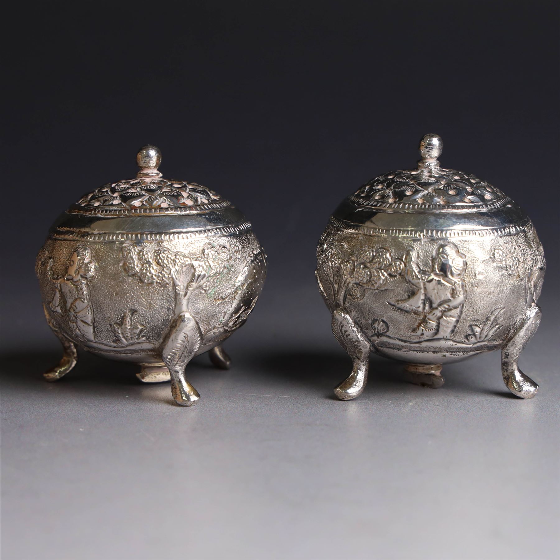 Pair of Middle Eastern silver cruets, of spherical form with pierced tops, embossed with figures, elephants and village scenes, unmarked, together with a pair of silver condiment spoons embossed with floral decoration 