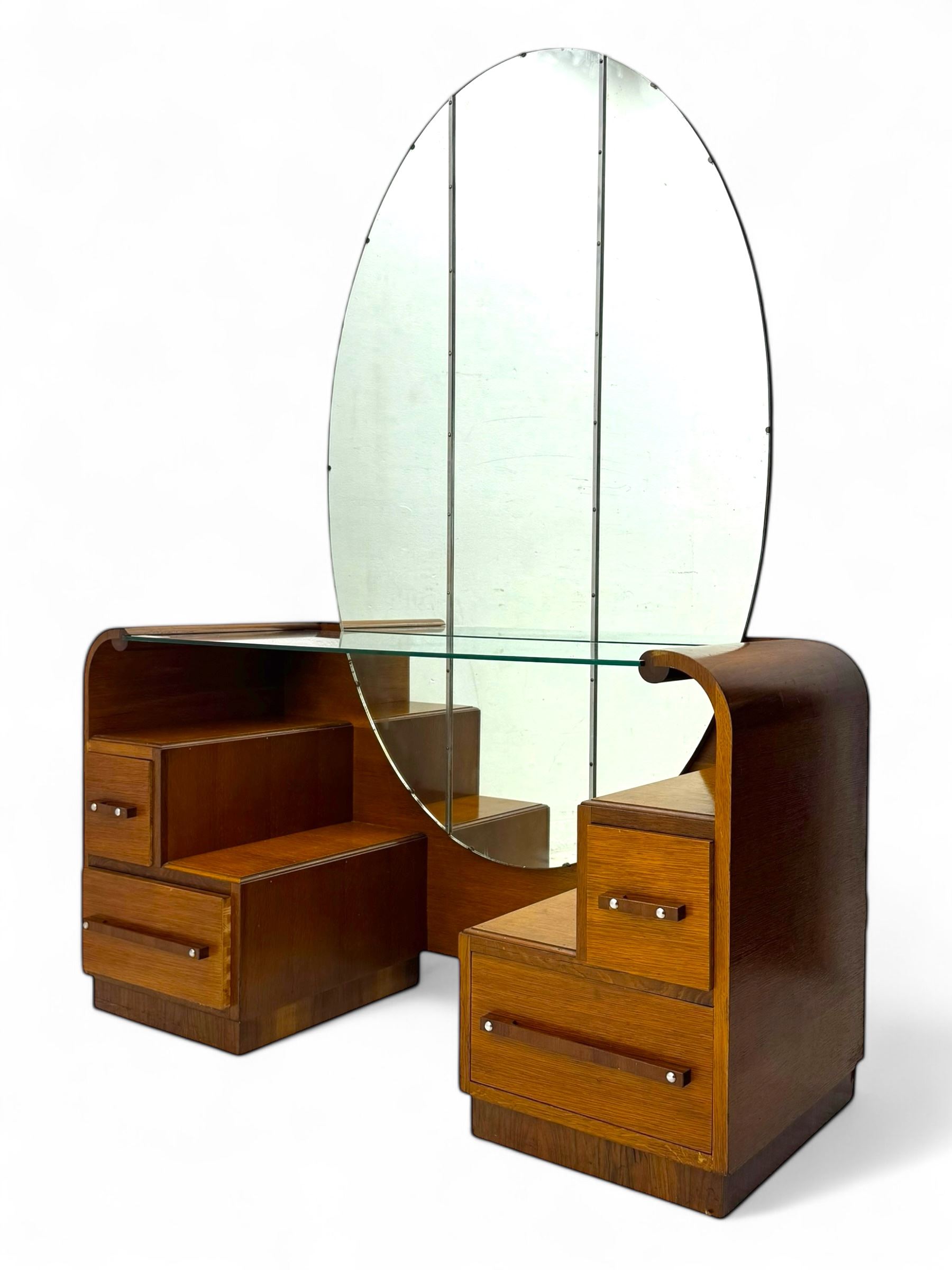 Early 20th century Art Deco walnut dressing table, featuring a large oval triptych mirror above a stepped base, fitted with four drawers and a central glass shelf, all raised on a plinth base