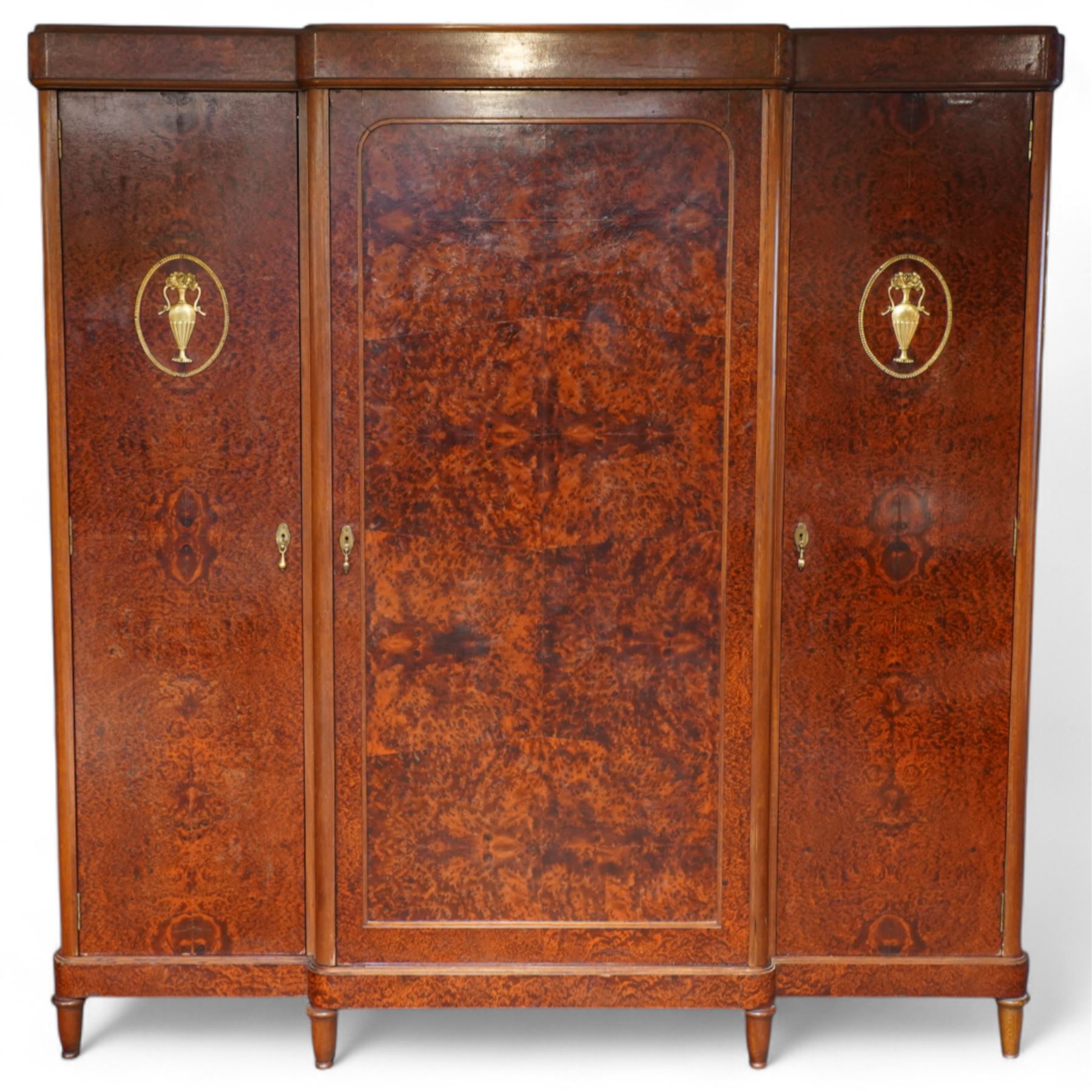 Early 20th century French burr maple breakfront armoire, enclosed by three panelled doors with bookmatched veneer and stringing, outer doors inlaid with gilt urn medallions, fitted interior with hanging spaces flanking a shelved compartment, raised on short tapered legs