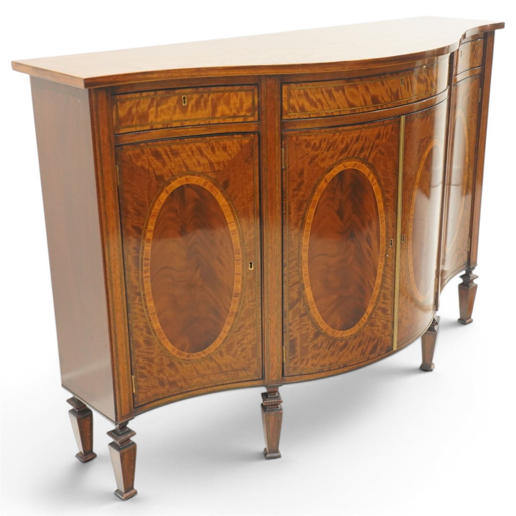 Quality handmade Sheraton Revival inlaid mahogany serpentine-front commode sideboard, shaped rectangular top with boxwood stringing, the banded frieze fitted with three drawers, over four matched-veneer cupboards with oval inlays and banding, raised on square tapering and turned feet with gadroon carving, retailed by David Hick Interiors, Jersey