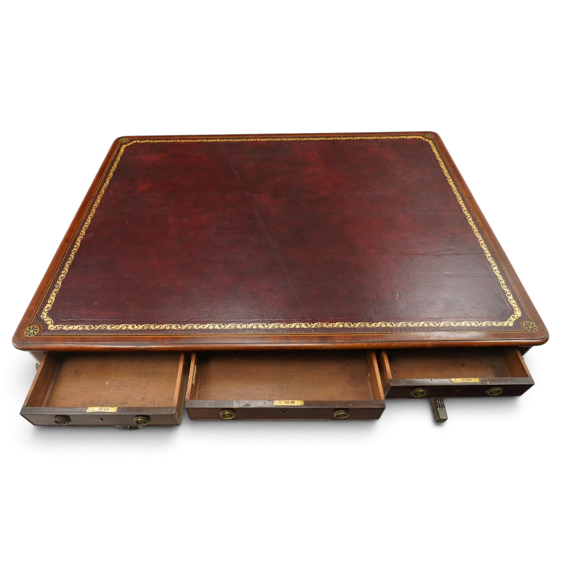 Regency inlaid mahogany and brass mounted library table, the rectangular top inset with gilt tooled burgundy leather writing surface, enclosed by  crossbanding and boxwood stringing with roundel corner motifs, above three cock-beaded frieze drawers to either side fitted with brass ring handles and brass escutcheons, the end friezes centred by applied brass foliate mounts, raised on twin reeded column supports over rectangular platform ends, united by a ring-turned stretcher, the reeded sabre legs terminating in brass caps and castors, one drawer bearing ivorine label 'Spillman & Co., St Martins Lane, London'