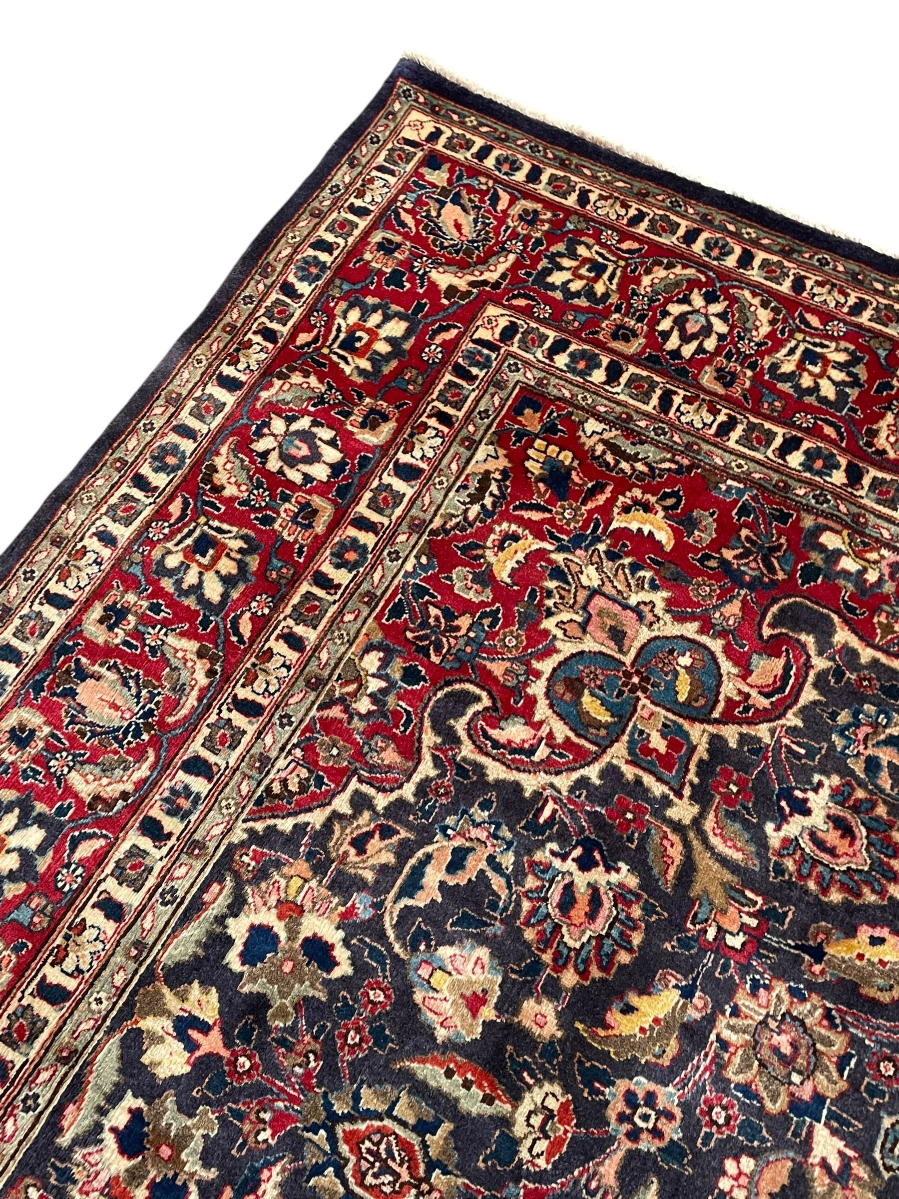 Persian Kashan indigo ground carpet, large central rosette medallion decorated with alternating palmettes, the surrounding field and spandrels with overall floral design depicting trailing branches and stylised plant motifs, crimson ground border with trailing floral pattern, enclosed within guard stripes 