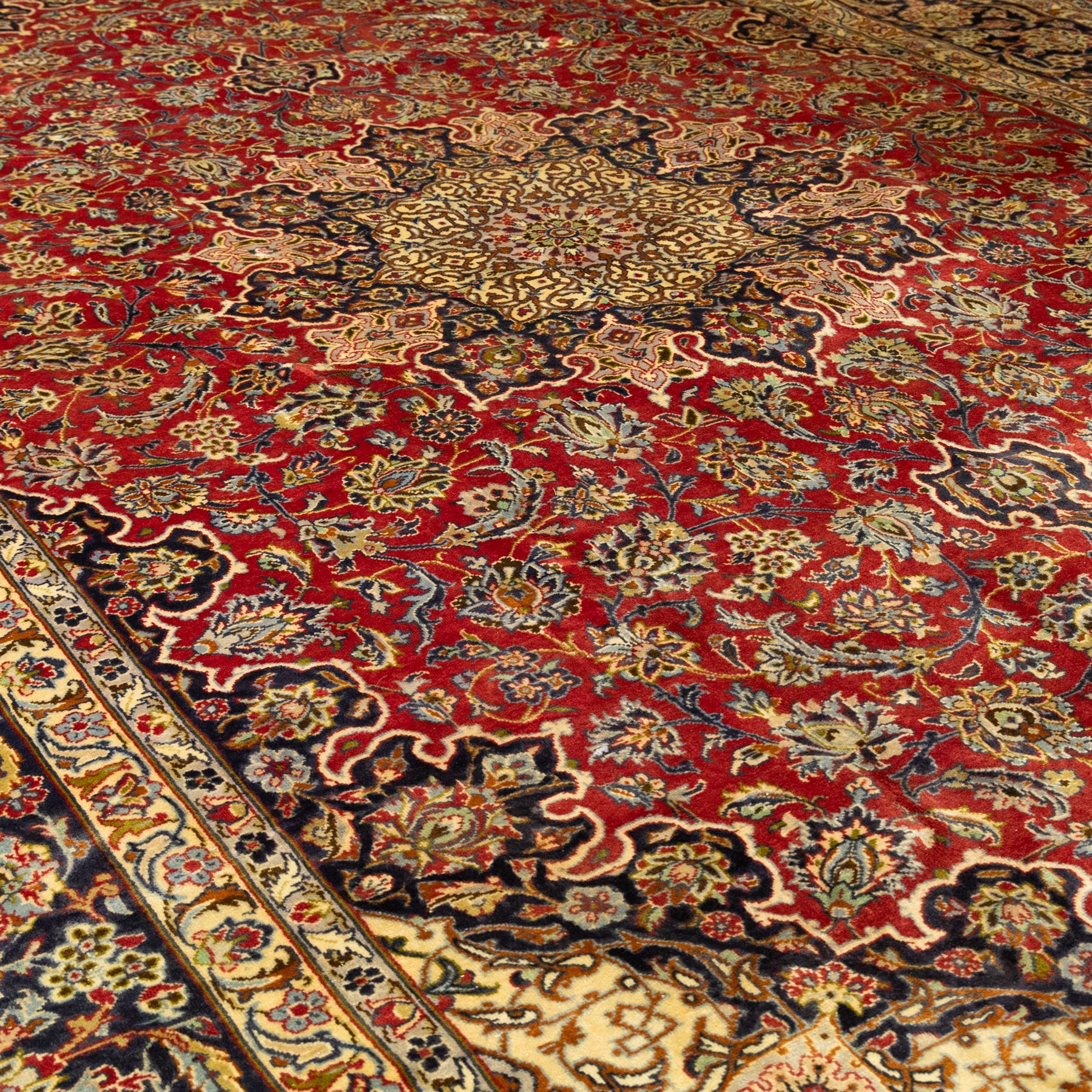 Central Persian Kashan hand-knotted carpet, crimson ground, with a large central ivory and dark navy floral medallion with pendants, surrounded by dense scrolling foliate and floral motifs incorporating rosettes, palmettes and vine work, enclosed by ivory spandrels and a dark navy border with repeating palmettes and meandering vines, flanked by floral guard stripes