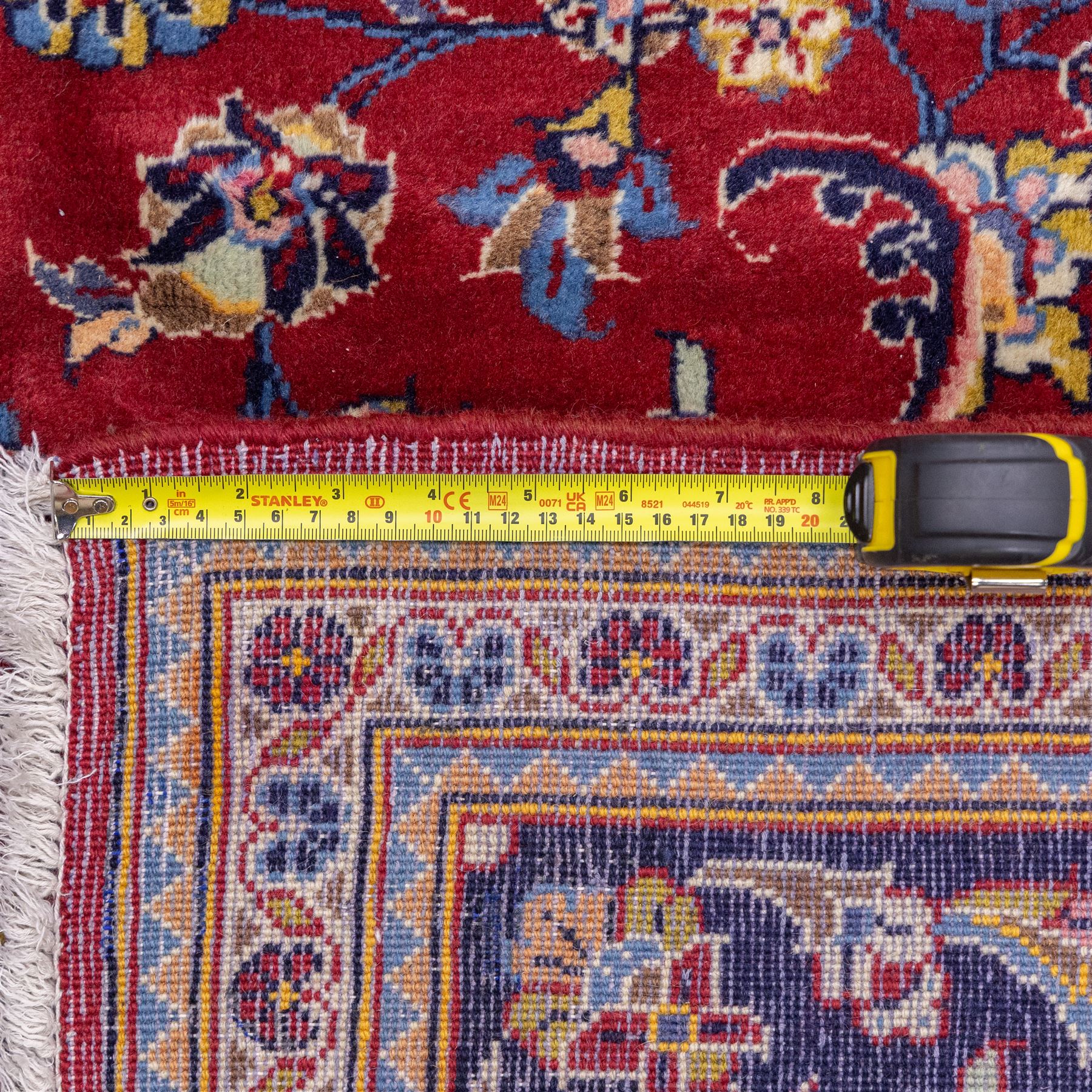 Central Persian Kashan hand-knotted rug, crimson field, decorated with an all-over floral and foliate design incorporating stylized palmettes, rosettes, and scrolling vine work, enclosed by a dark navy blue main border ornamented with flowering branches and palmettes flanked by multiple narrow guard borders with repeating floral motifs, with cotton fringe ends 