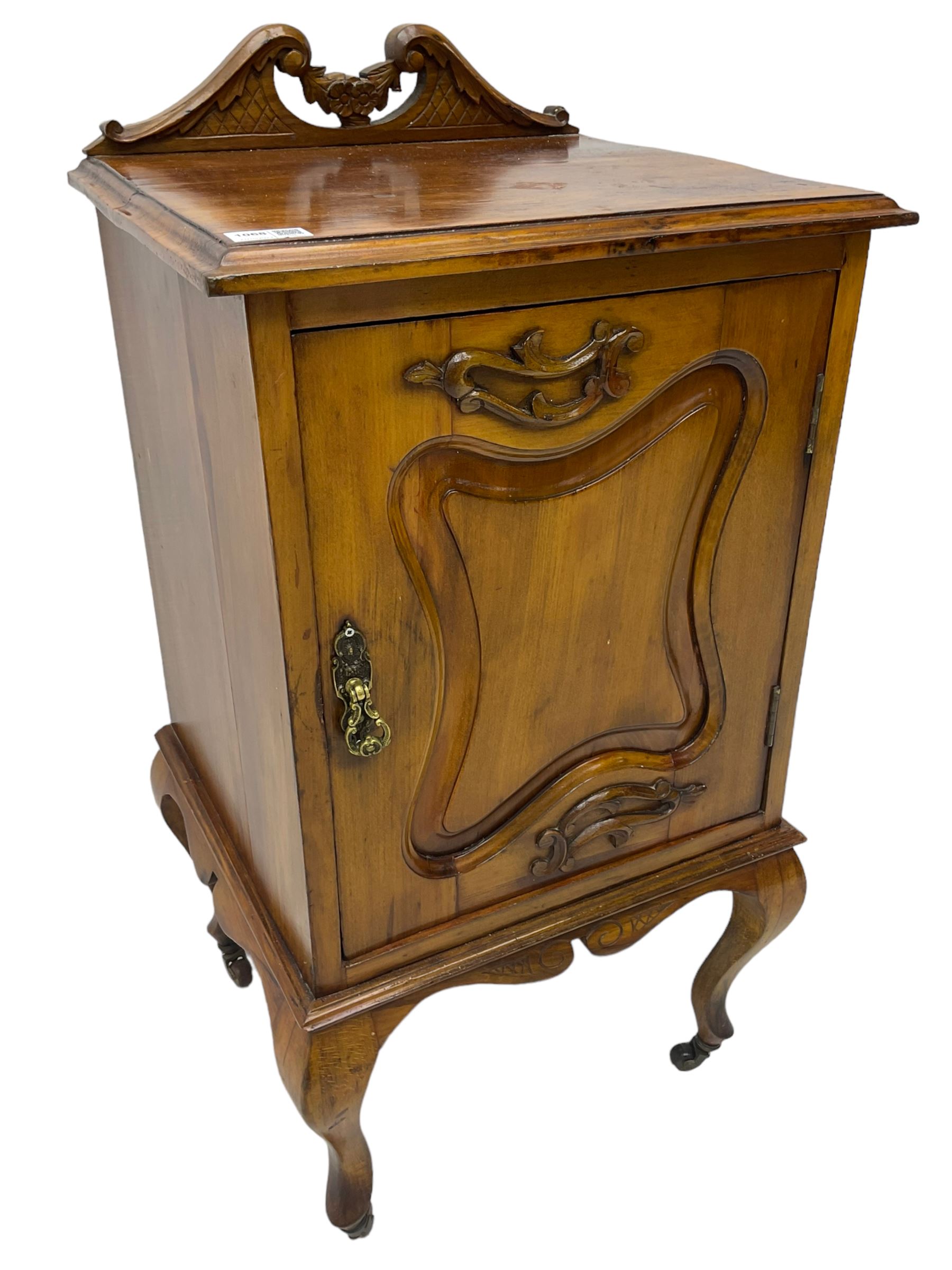 Edwardian Art Nouveau walnut bedside cabinet, raised broken swan neck back with carved flower heads, single fielded panelled door enclosing single shelf, raised on cabriole supports with ceramic castors