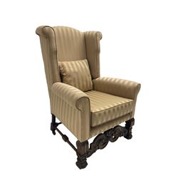 William and Mary style wingback armchair, upholstered in gold striped fabric, on walnut scroll carved supports joined by scroll and foliate carved middle rail, turned H-shaped stretchers, on castors