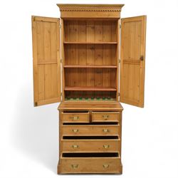 Early 20th-century pine gun cabinet with later fitted shelves, of two-part construction, upper cupboard moulded and dentil cornice above two panelled doors revealing three removable shelves and circular recesses for gun barrels at the top, lower section with two short and three long graduated drawers with brass swan neck handles on plinth base 