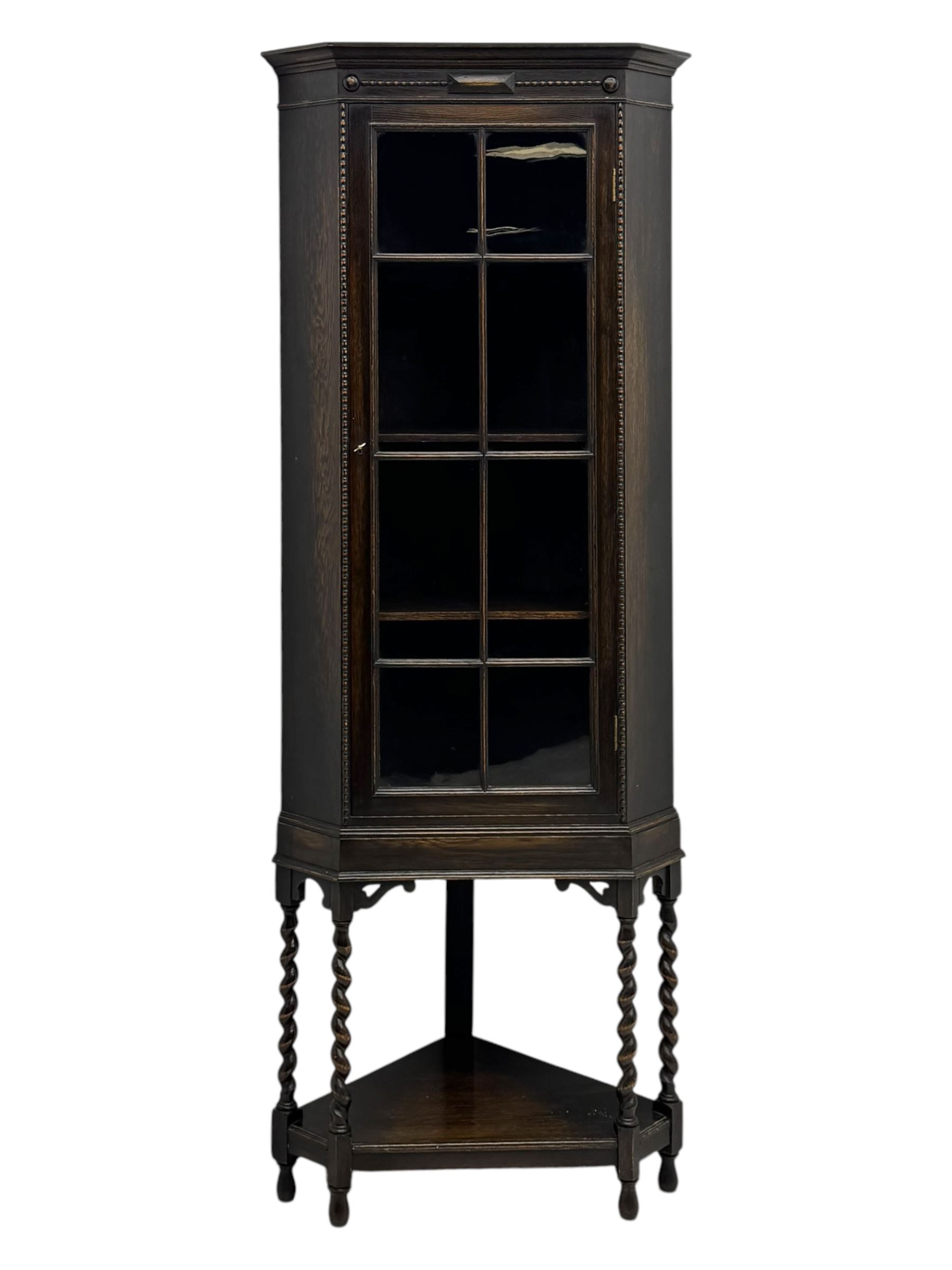 Early 20th century oak corner display cabinet, the moulded canted cornice over a single glazed door with geometric astragal bars, shelved interior, the lower tier forming a triangular platform, raised on four barley-twist supports united by shaped brackets