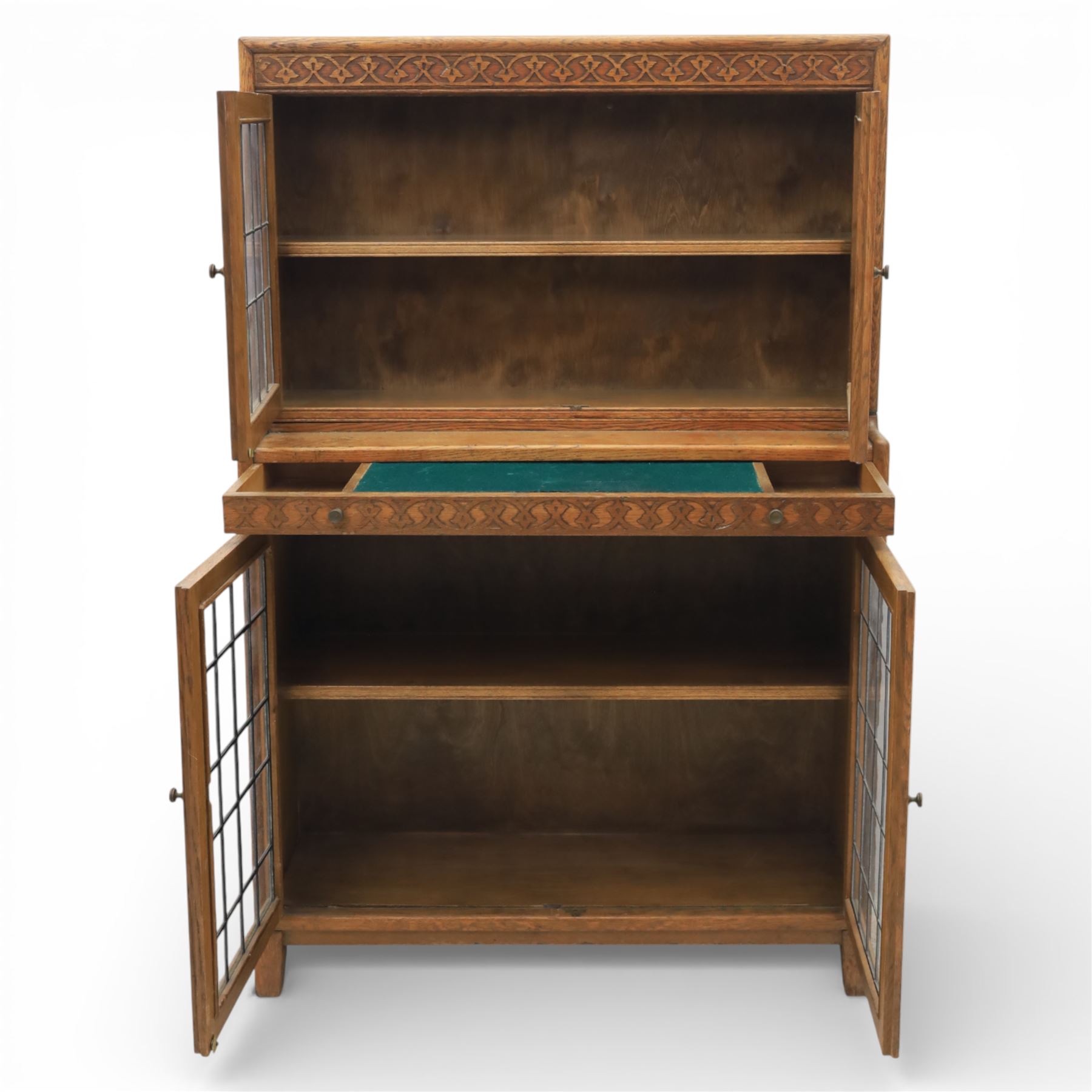 Early 20th-century Arts and Crafts glazed oak bookcase, upper section fitted with a pair of leaded glazed doors enclosing a fixed shelf above a projecting frieze drawer interior with baize-lined writing surface flanked by two compartments, carved with interlaced decoration, lower section fitted with a further pair of leaded glazed doors enclosing a single shelf, raised on short stile feet 
