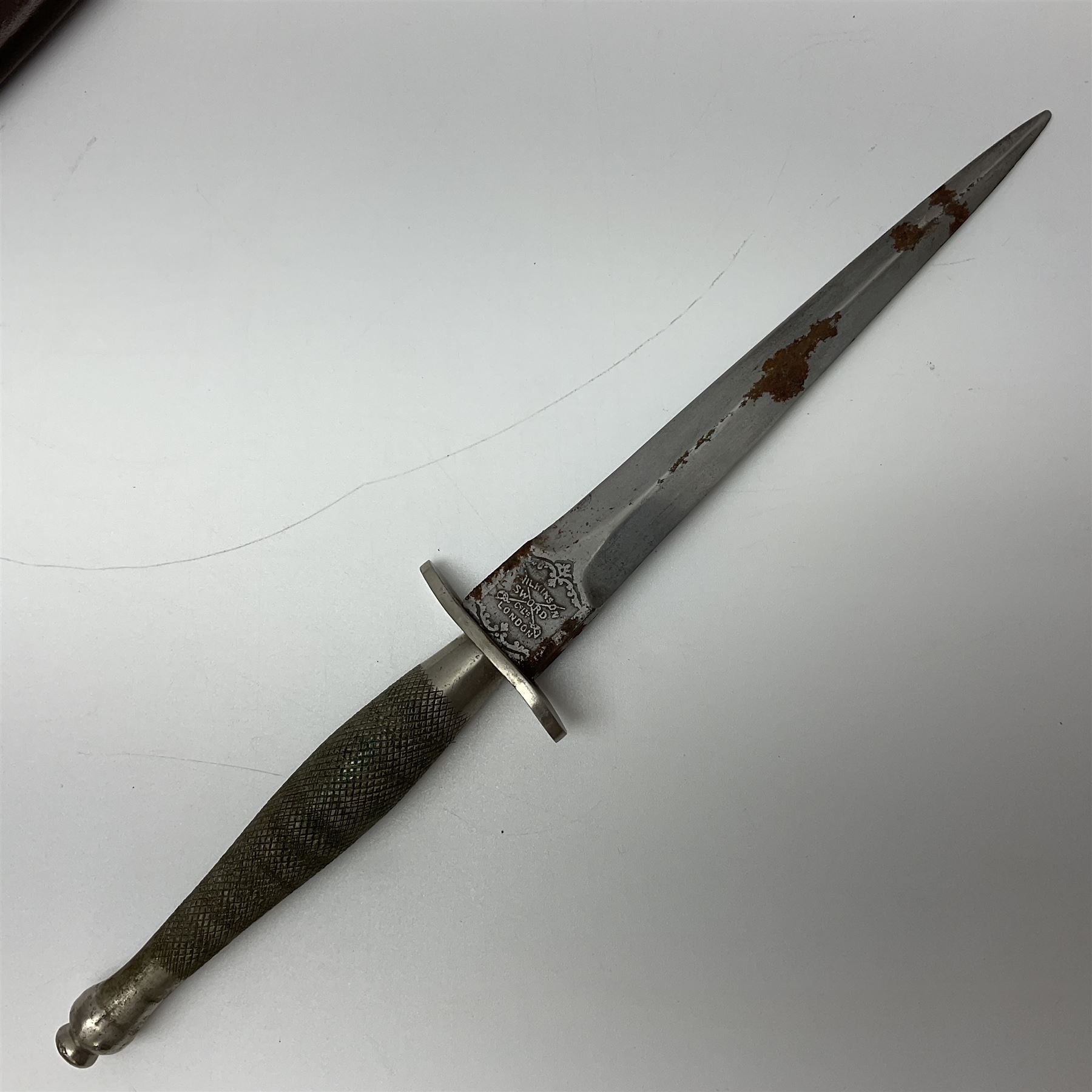 First Pattern Fairburn Sykes ''F-S'' Fighting Knife by Wilkinson Sword Ltd., the 17cm double edge steel blade with raised medial ridge to each side, each side of the ricasso with a recessed panel, one side with maker's name, the opposing side etched THE F~S FIGHTING KNIFE, with nickel plated brass recurving crossguard and diamond cut grip; in leather scabbard with metal chape