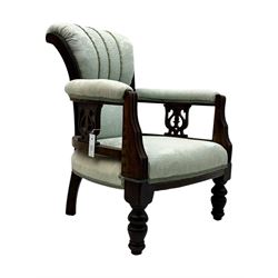 Late Victorian carved mahogany open armchair, the shaped back with fluted upholstered panel and padded arms above pierced scroll carved side supports, over a broad upholstered seat, on turned front supports