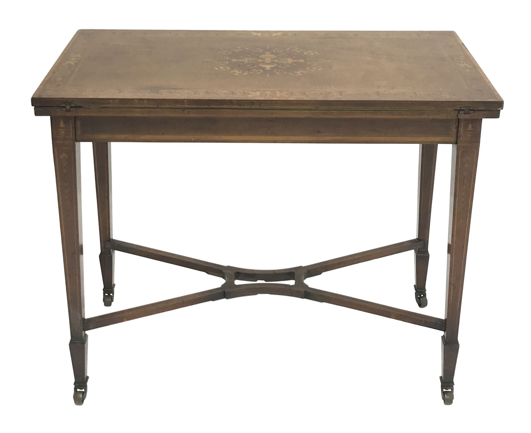 Edwardian walnut and mahogany Sheraton revival card table, rectangular top cross banded and inlaid with scrolling foliage, the central inlay with four urns and trailing leafage, swivel fold over action revealing storage well and baize lined interior, husk inlaid square tapering supports with spade feet, shaped and pierced x-framed stretcher, brass castors