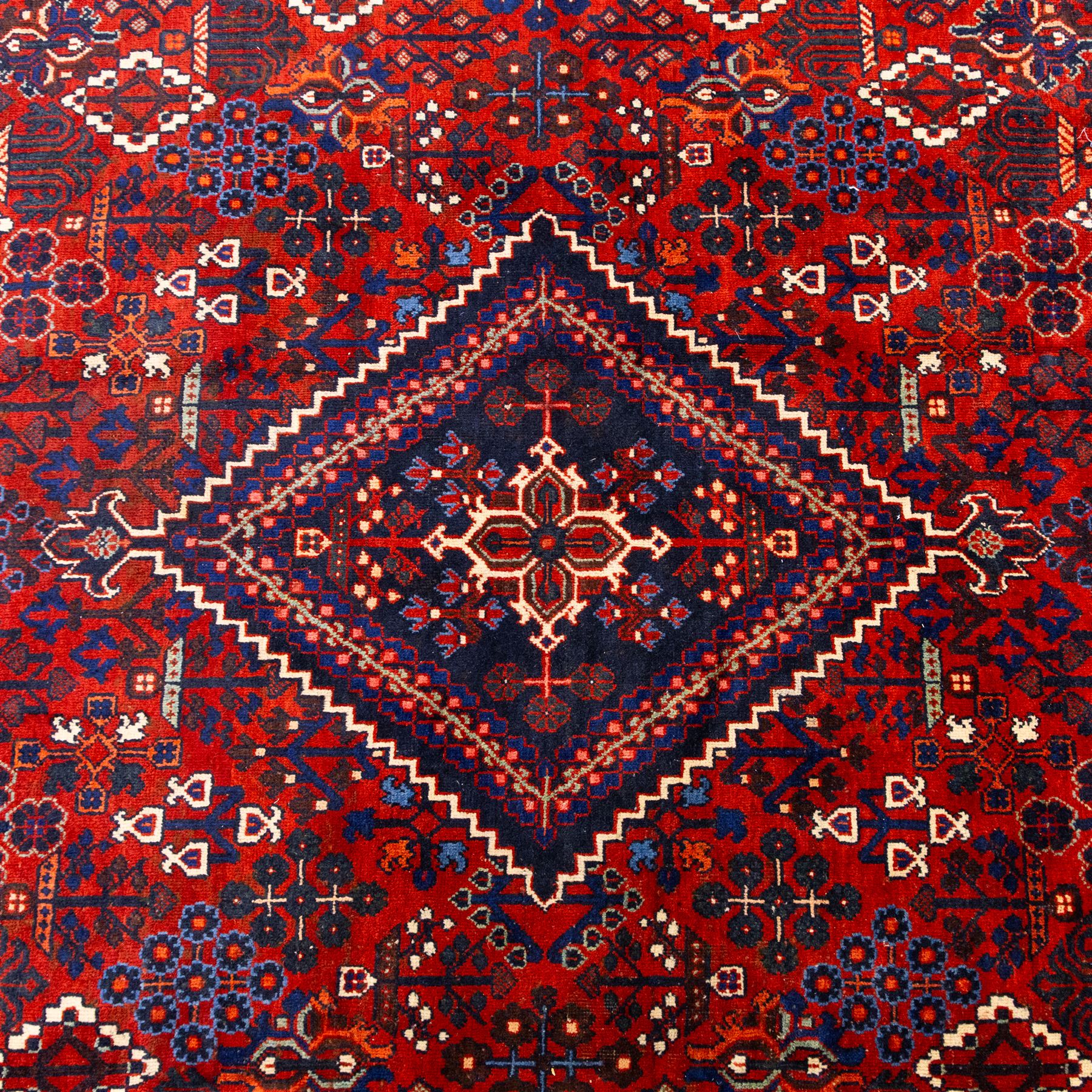 Central Persian Joshagen hand-knotted carpet, madder-red field with a central stepped lozenge medallion and densely filled with stylized geometric floral and tree motifs arranged in a repeating lattice enclosed by indigo spandrels and a broad navy border of angular palmette and vine ornament, flanked by multiple guard stripes, cotton fringe ends 
