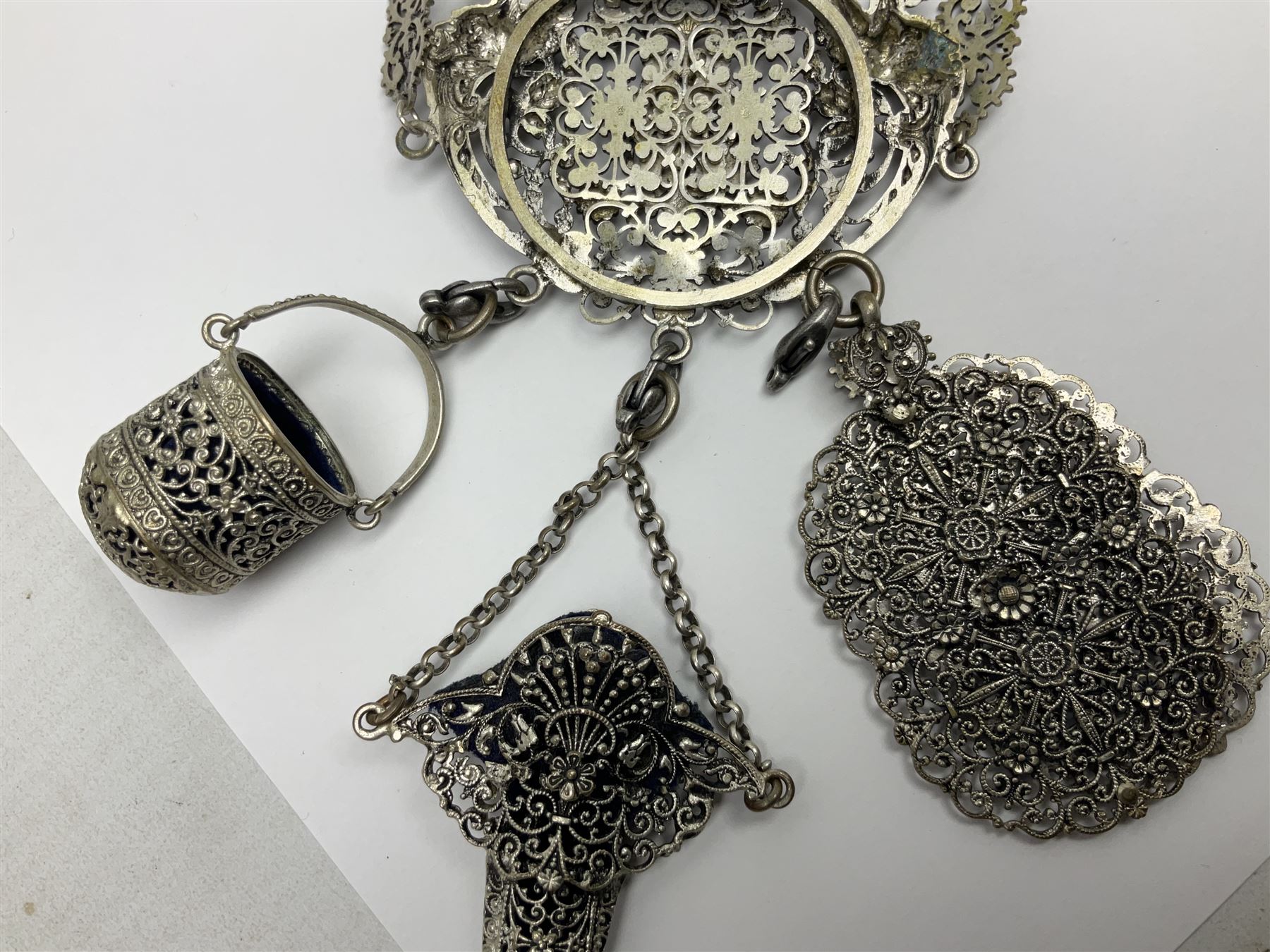 19th century continental silver plated chatelaine, the main body flanked with winged mythical beasts and central soldier slaying a dragon supporting three suspended items to include velvet lined small basket and long clasped filigree chain, together with a thimble