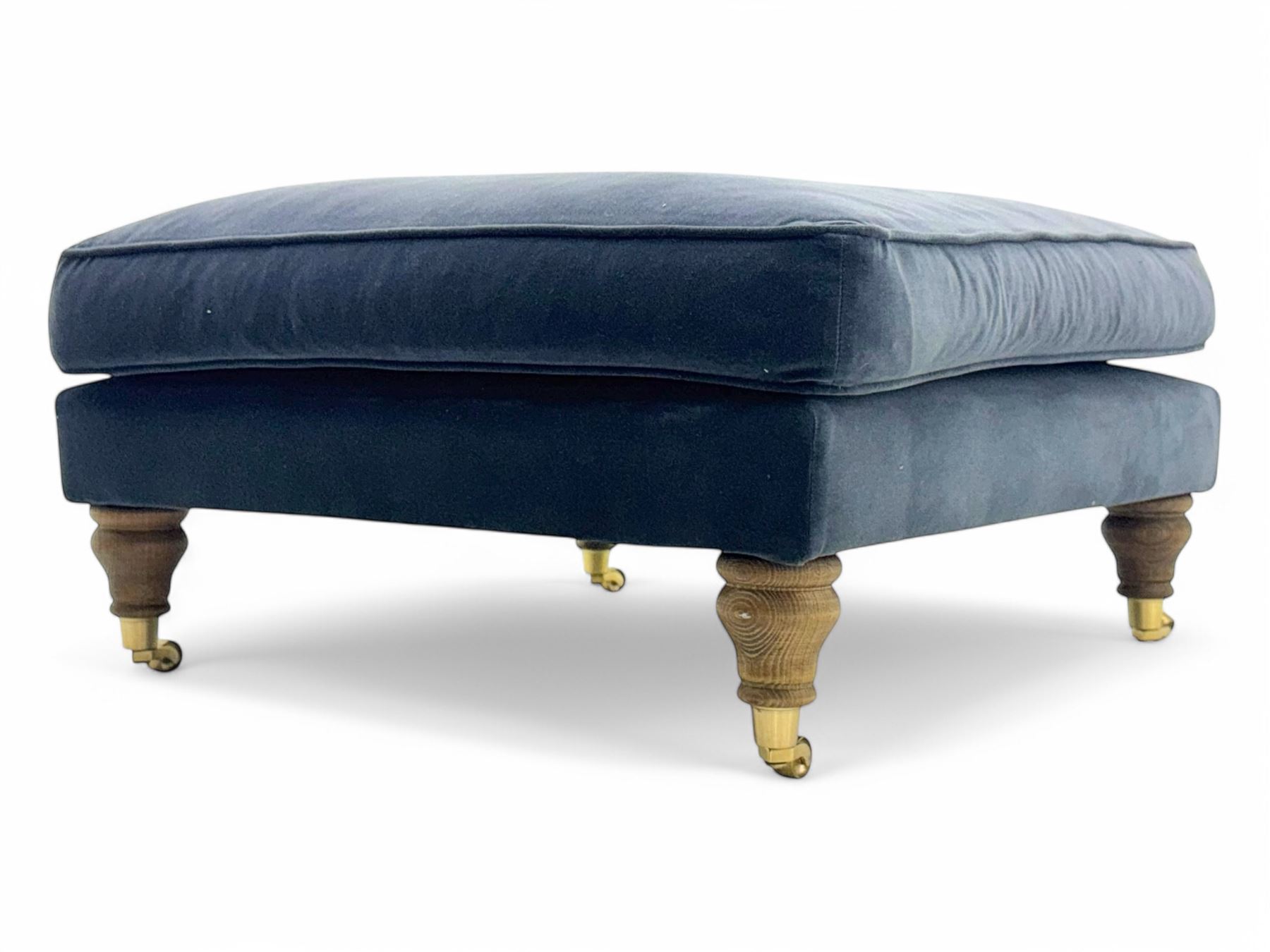 Contemporary square footstool upholstered in navy armour smart velvet, featuring a deep padded top, turned wood supports, and brass castors