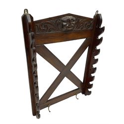 Victorian oak wall hanging whip or stick rack, sloped arch pediment carved with central grotesque mask with extended scrolling foliage, the upright supports cut with rests, x-framed back, two lower hooks