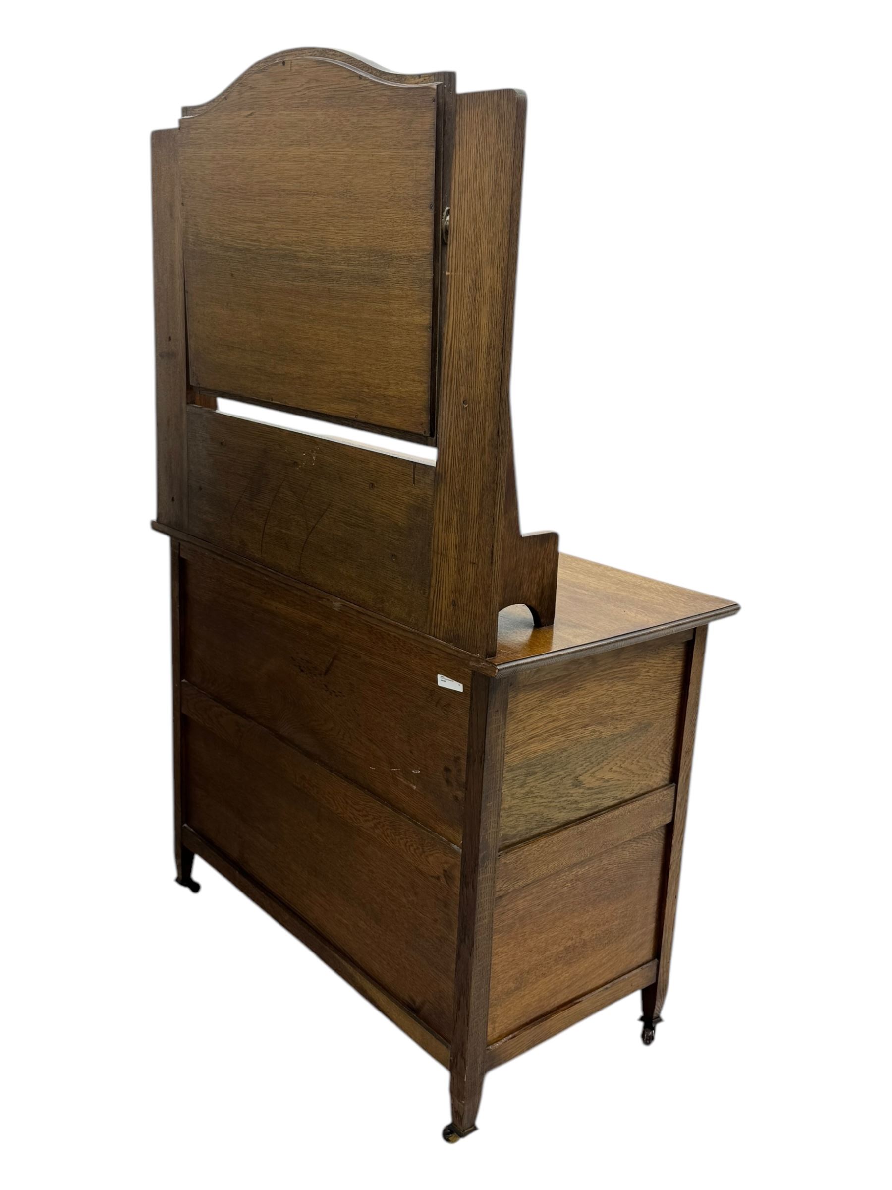 Early 20th century Arts and Crafts oak dressing chest, the arched mirror flanked by floral marquetry inlaid uprights above a shelf and drawer, over three long drawers with square hammered metal plates and circular pulls, on slender supports with castors